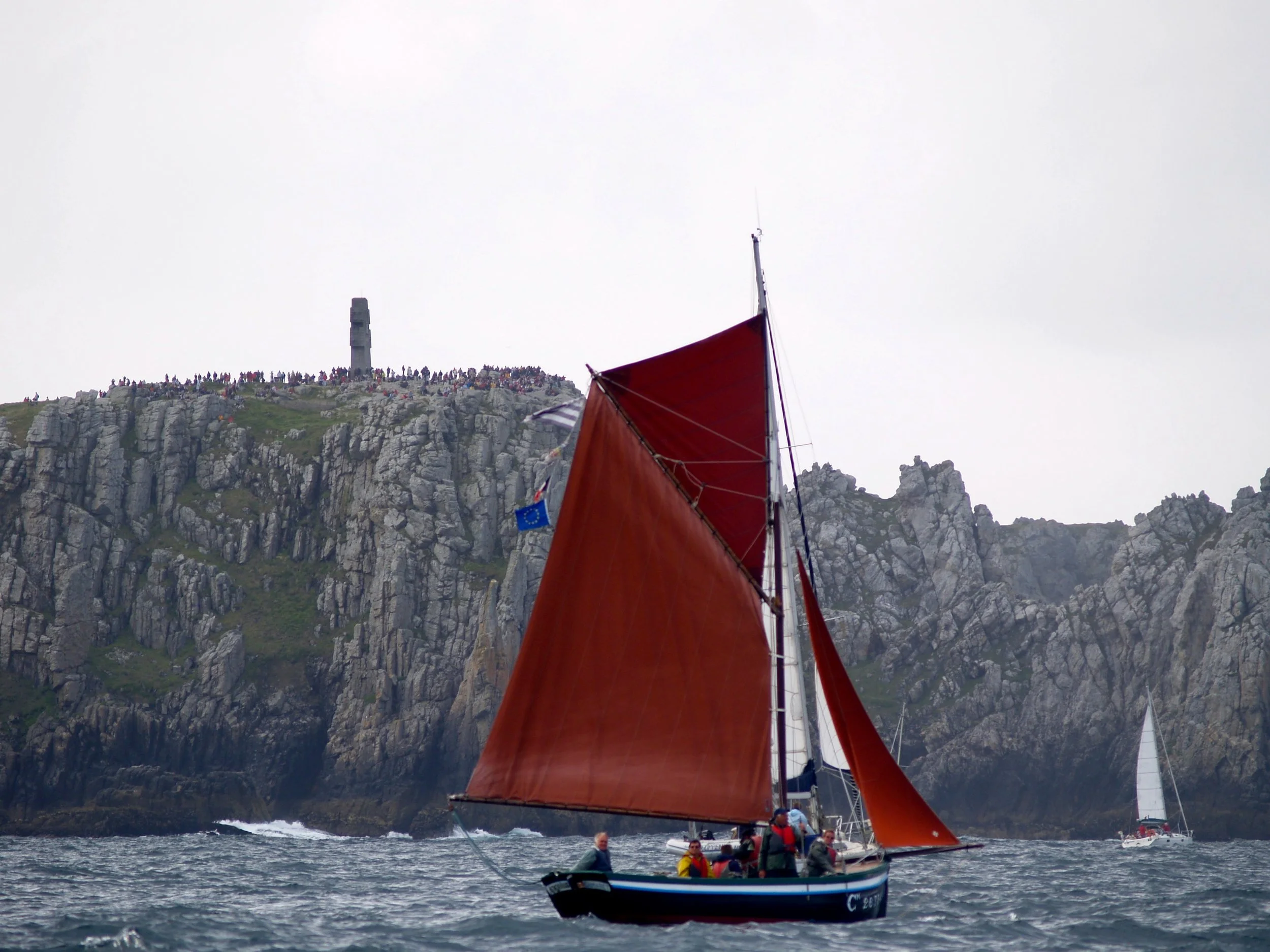 Un voilier avec une voile rouge navigue en mer, avec un groupe de personnes à bord, devant des falaises escarpées et un monument en haut de la falaise, par temps nuageux.