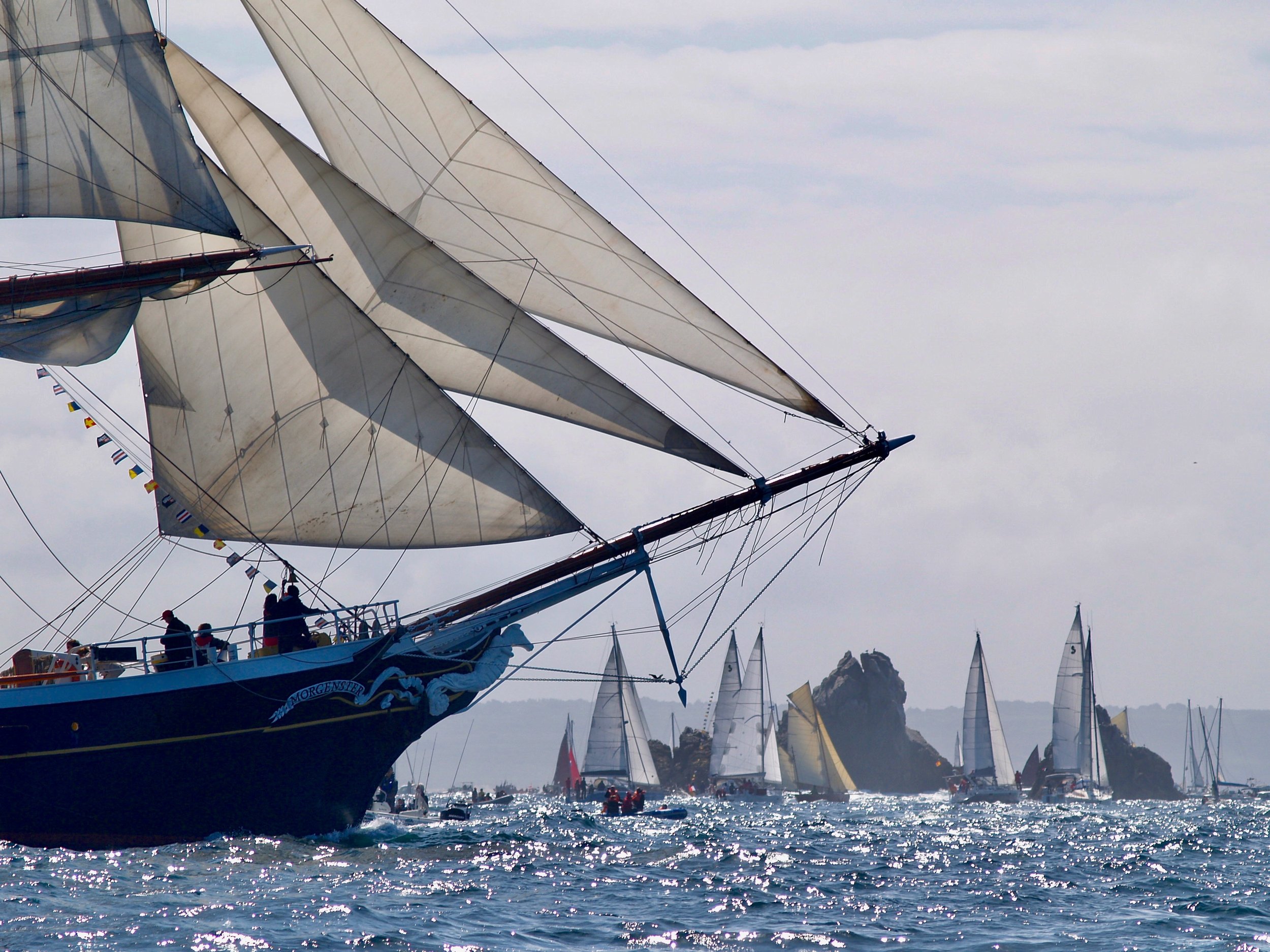 Une flotte de voiliers navigue en mer lors d'une régate. Le soleil brille, et le ciel est légèrement nuageux.