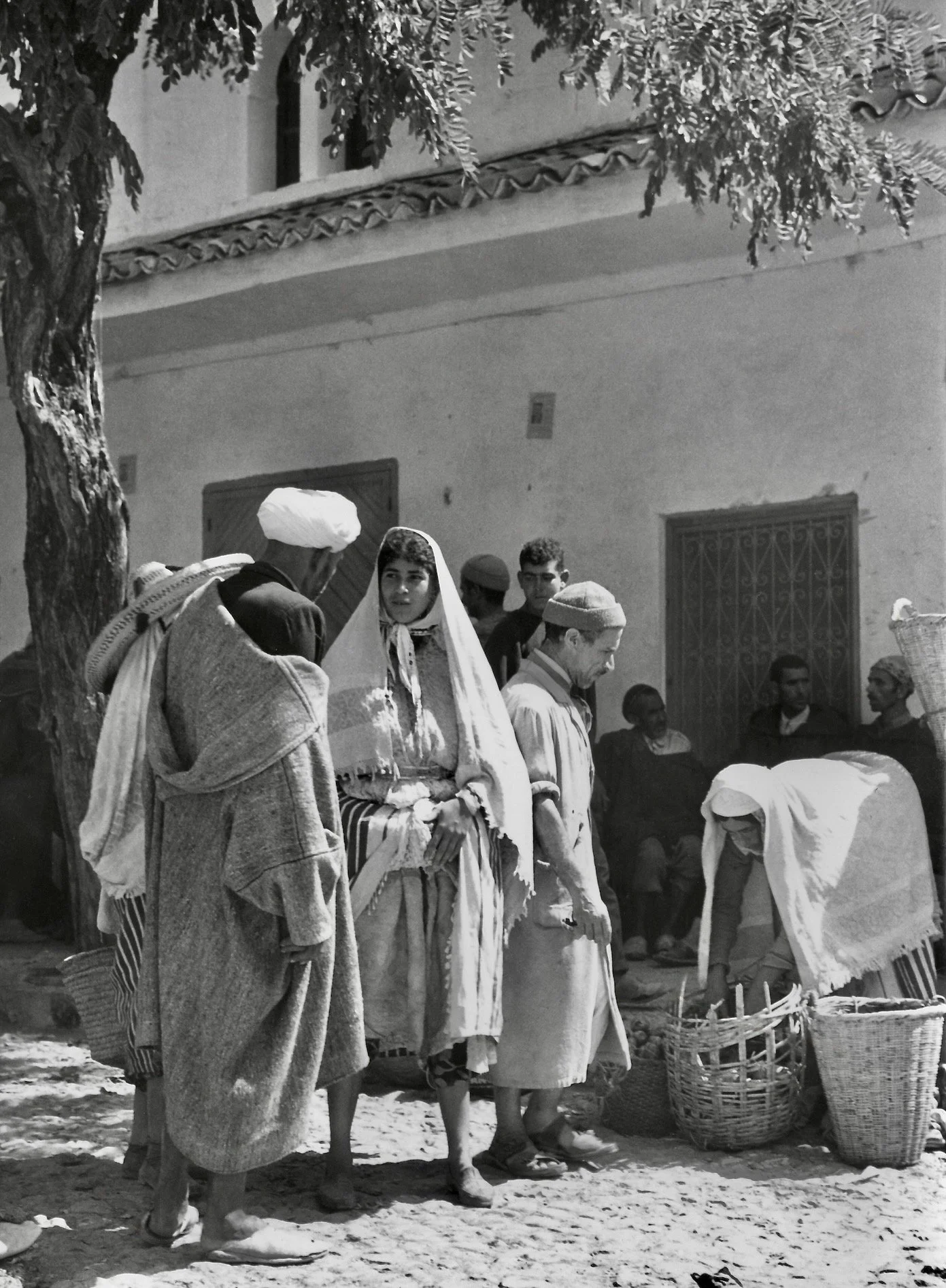 Groupe de personnes en vêtements traditionnels, certains portant des turbans, dans une scène en plein air avec des paniers en osier et un bâtiment en arrière-plan.