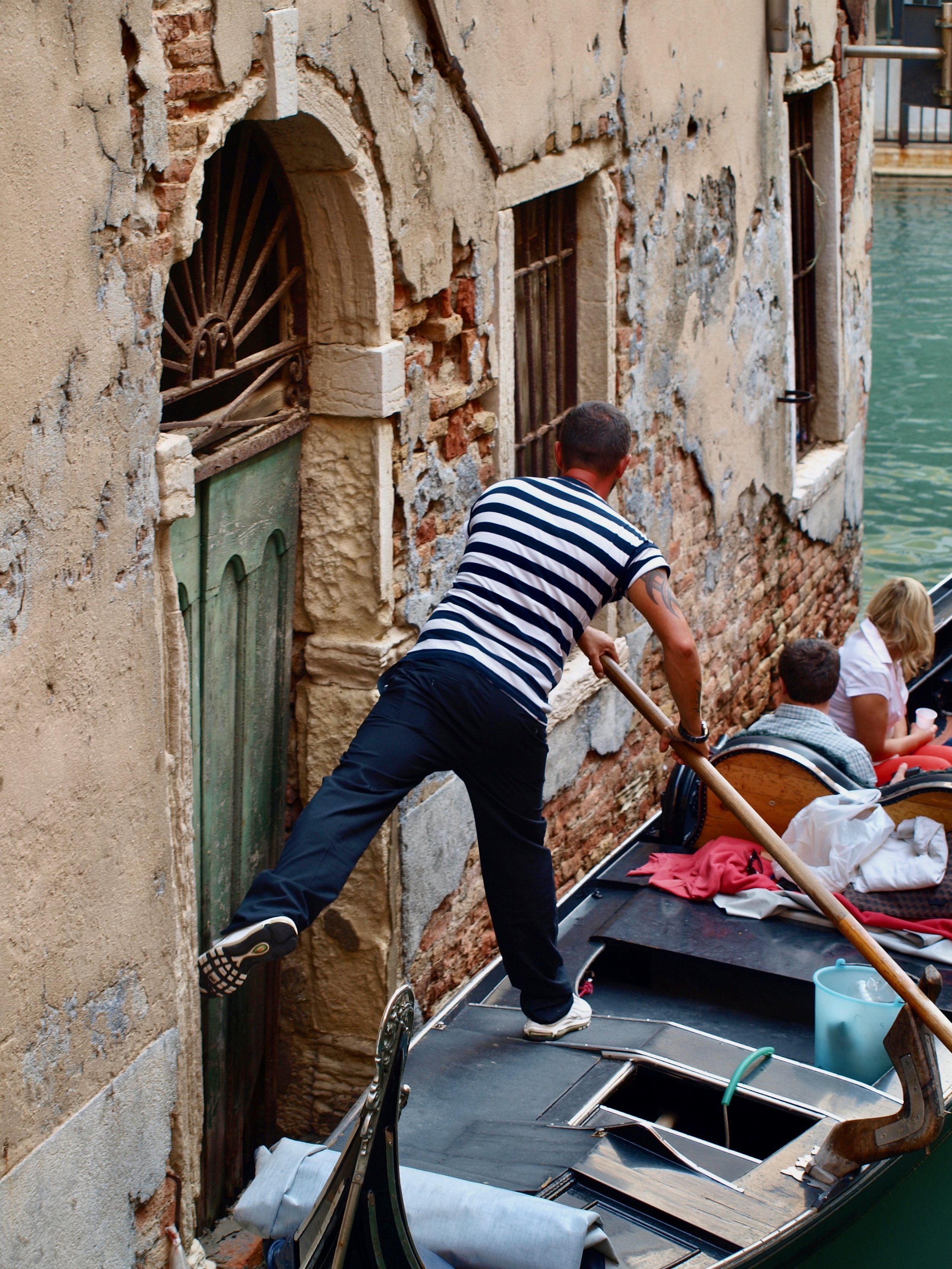 Un homme en t-shirt rayé noir et blanc pousse un bateau dans une rue avec un bâtiment en pierre endommagé. Il y a d'autres personnes dans le bateau, et des objets comme des vêtements et un seau visibles dans le bateau.