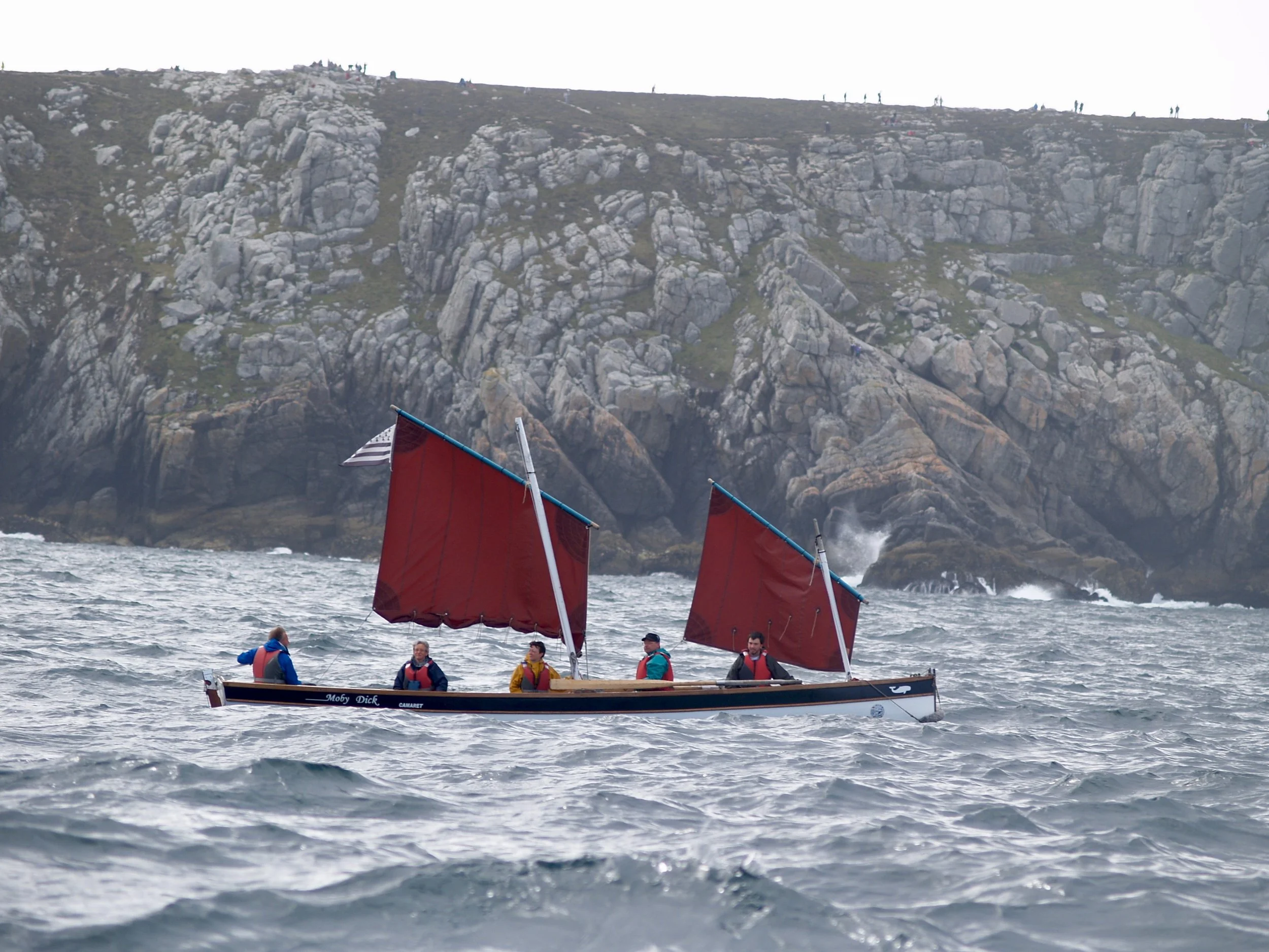 Un groupe de personnes naviguant sur un bateau à voile dans l'océan, avec des montagnes rocheuses en arrière-plan.