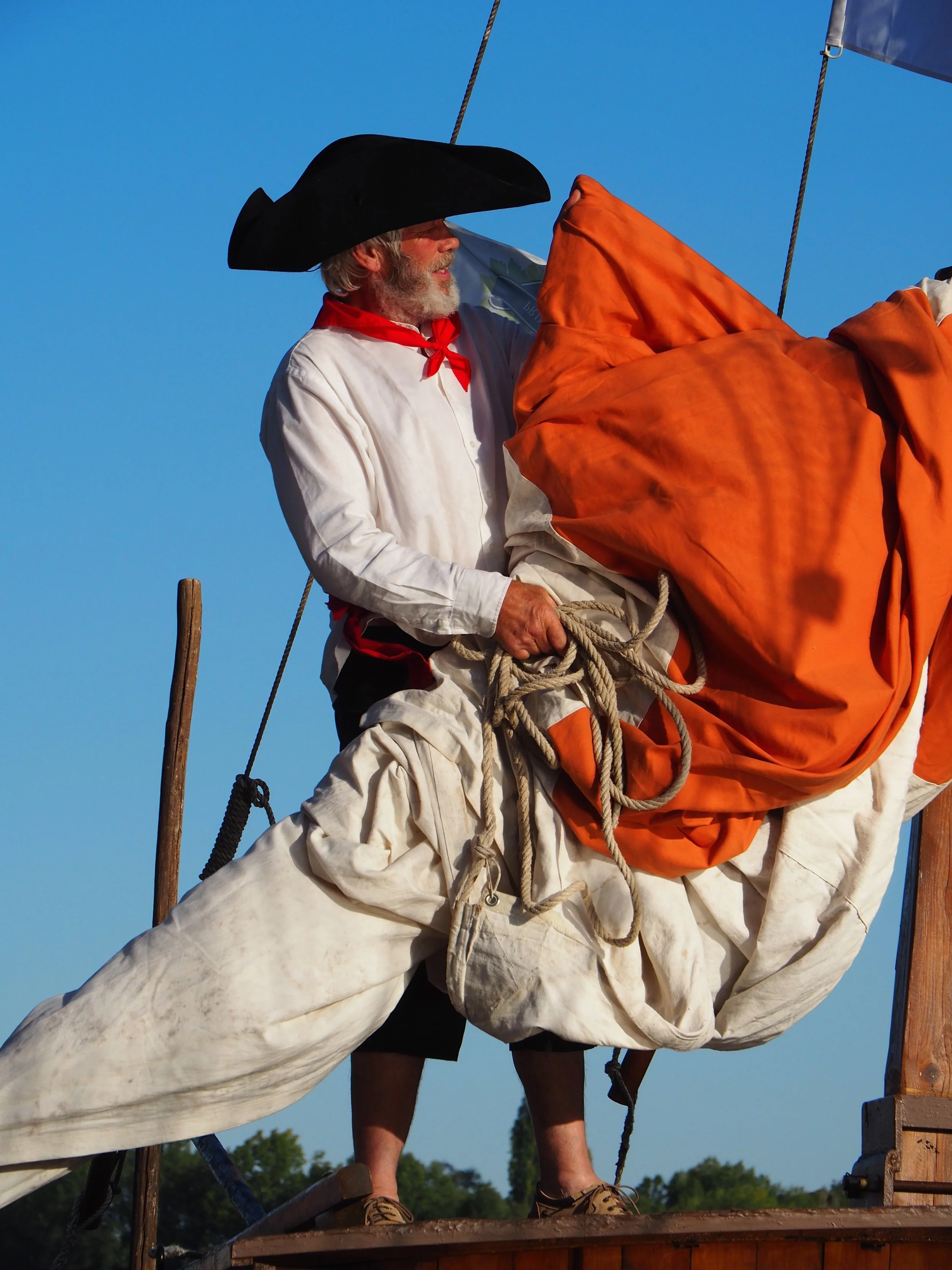 Une personne en costume historique avec un chapeau noir, habillée en blanc avec une écharpe rouge, debout sur un bateau, tenant une corde, sous un ciel bleu.