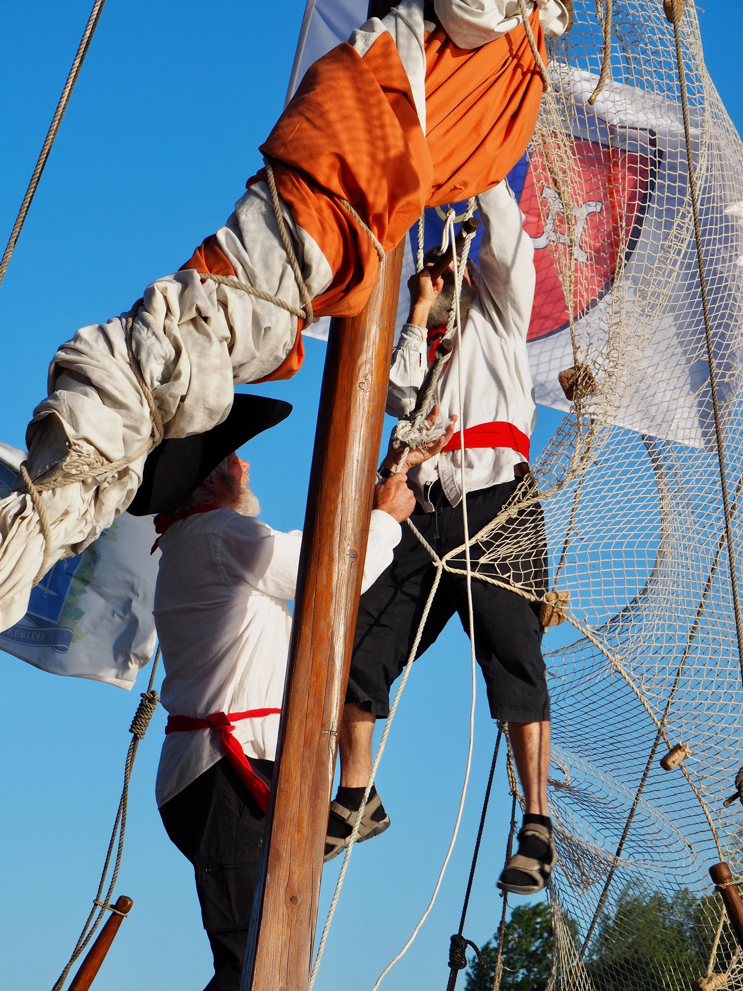 Des personnes habillées de costumes d'époque participent à une reconstitution historique sur un navire, en montant ou en travaillant sur la mâture contre un ciel bleu clair.