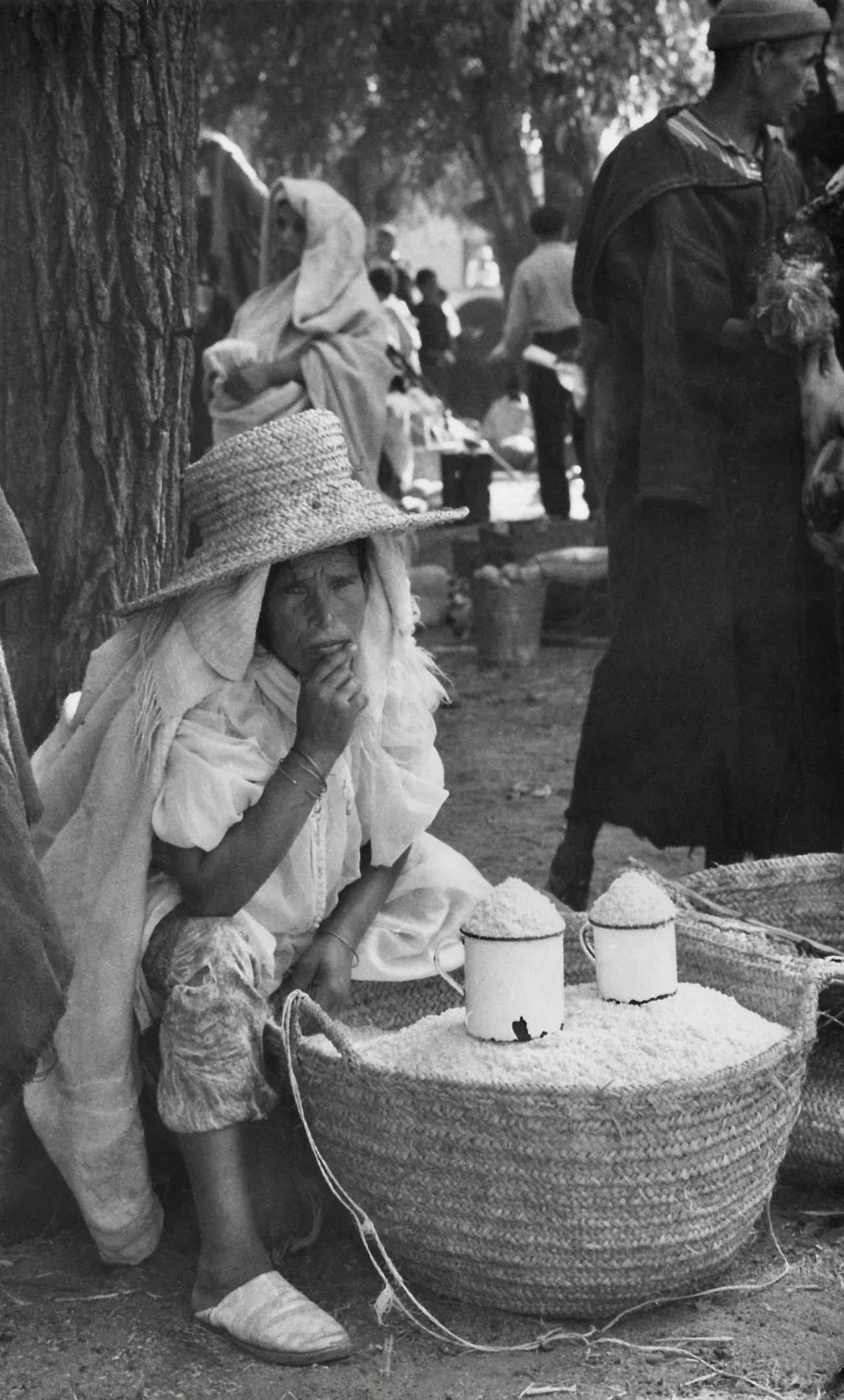 Femme assise, portant un chapeau en paille, fume une cigarette, devant un grand panier en osier rempli de riz, dans un marché en plein air. Autres personnes en arrière-plan, certaines vendant ou achetant des marchandises, arbres et lumière naturelle.