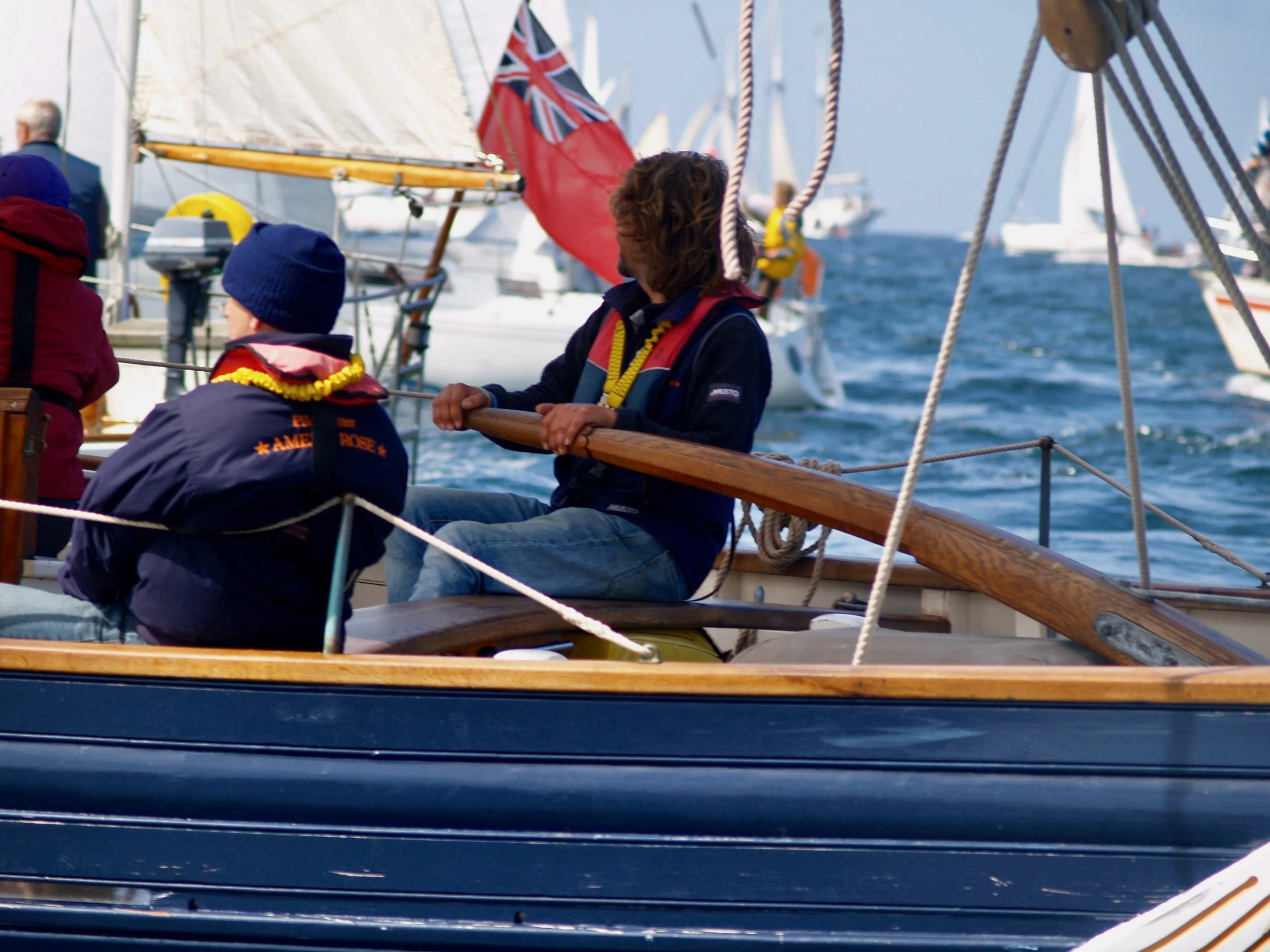 Jeunes sur un bateau à voile avec des vestes de sécurité, regardant vers l'horizon où se trouvent d'autres voiliers en mer, drapeaux britanniques flottant sur le bateau.