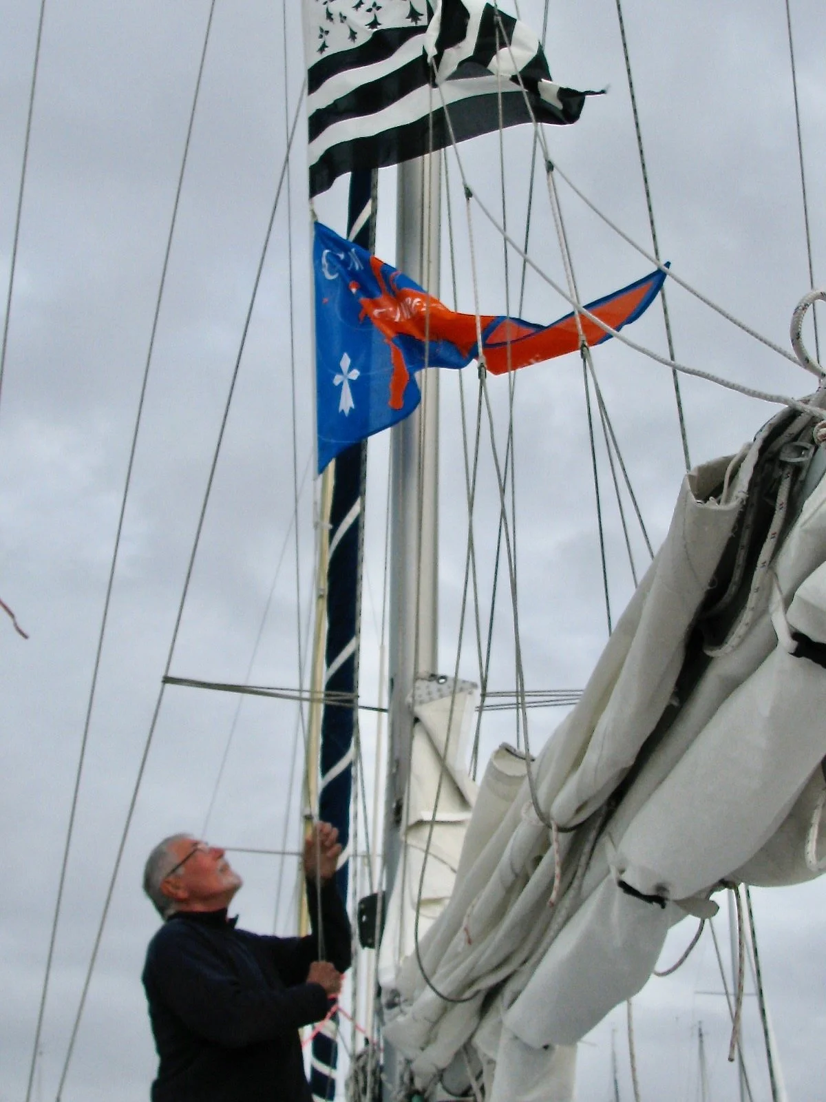Une personne âgée en costume noir ajuste une voile sur un mât de bateau en mer par jour nuageux.