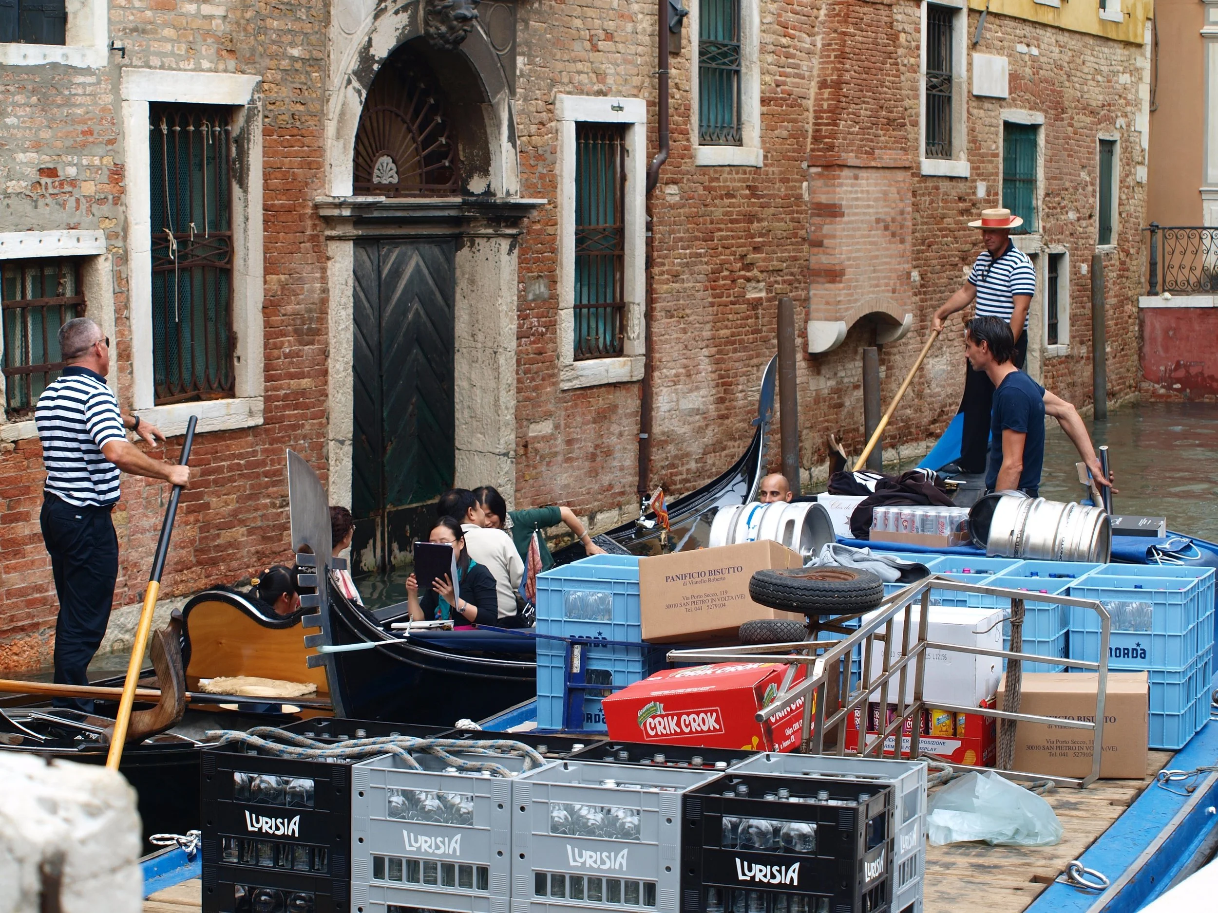 Gondole avec des personnes naviguant dans un canal à Venise, entourée de bâtiments en brique, avec des boîtes en plastique et des accessoires de bateau sur le quai.