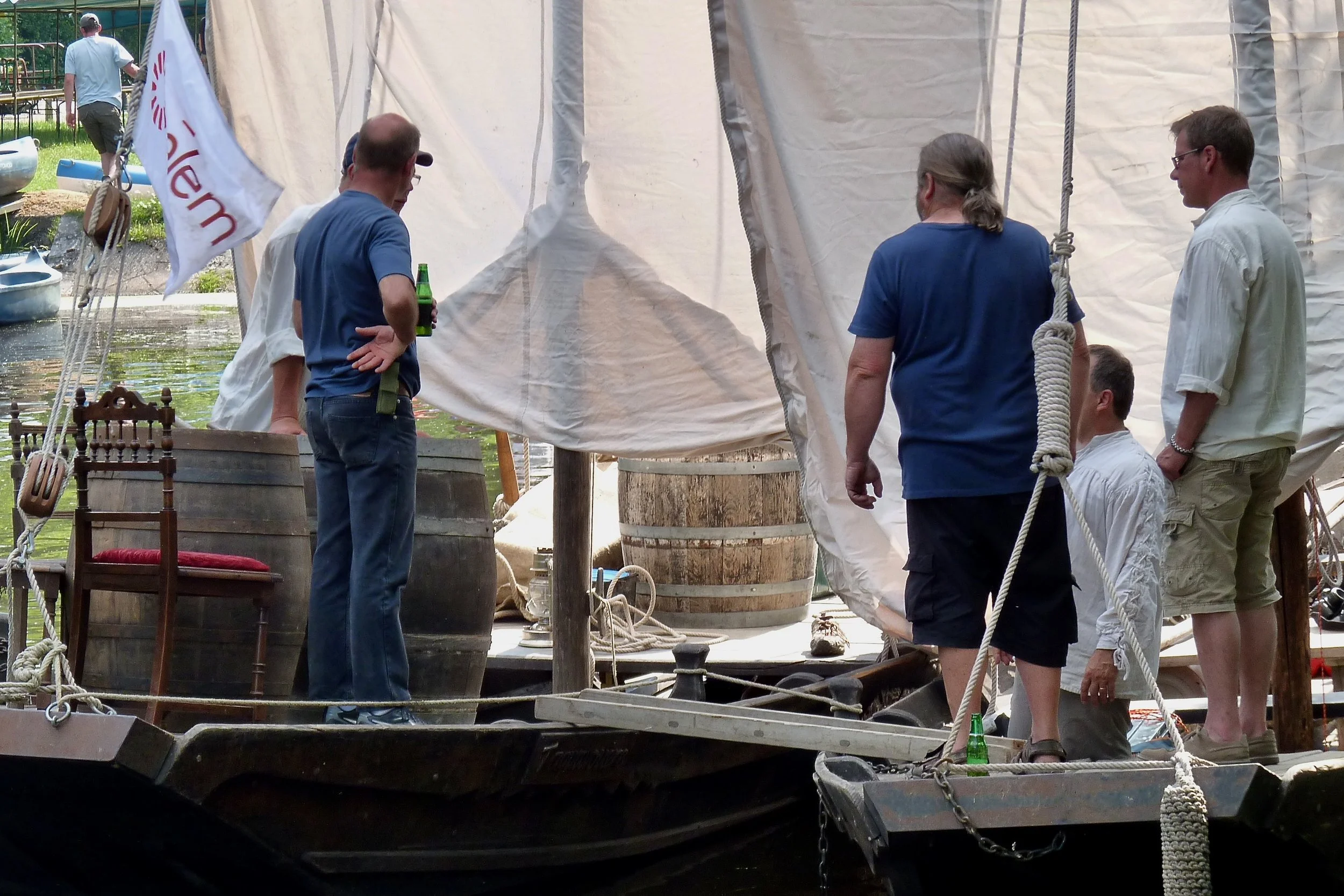 Groupe de personnes discutant sur un bateau en bois, avec des tonneaux en bois, une bannière, et des bouteilles vertes sur le pont.