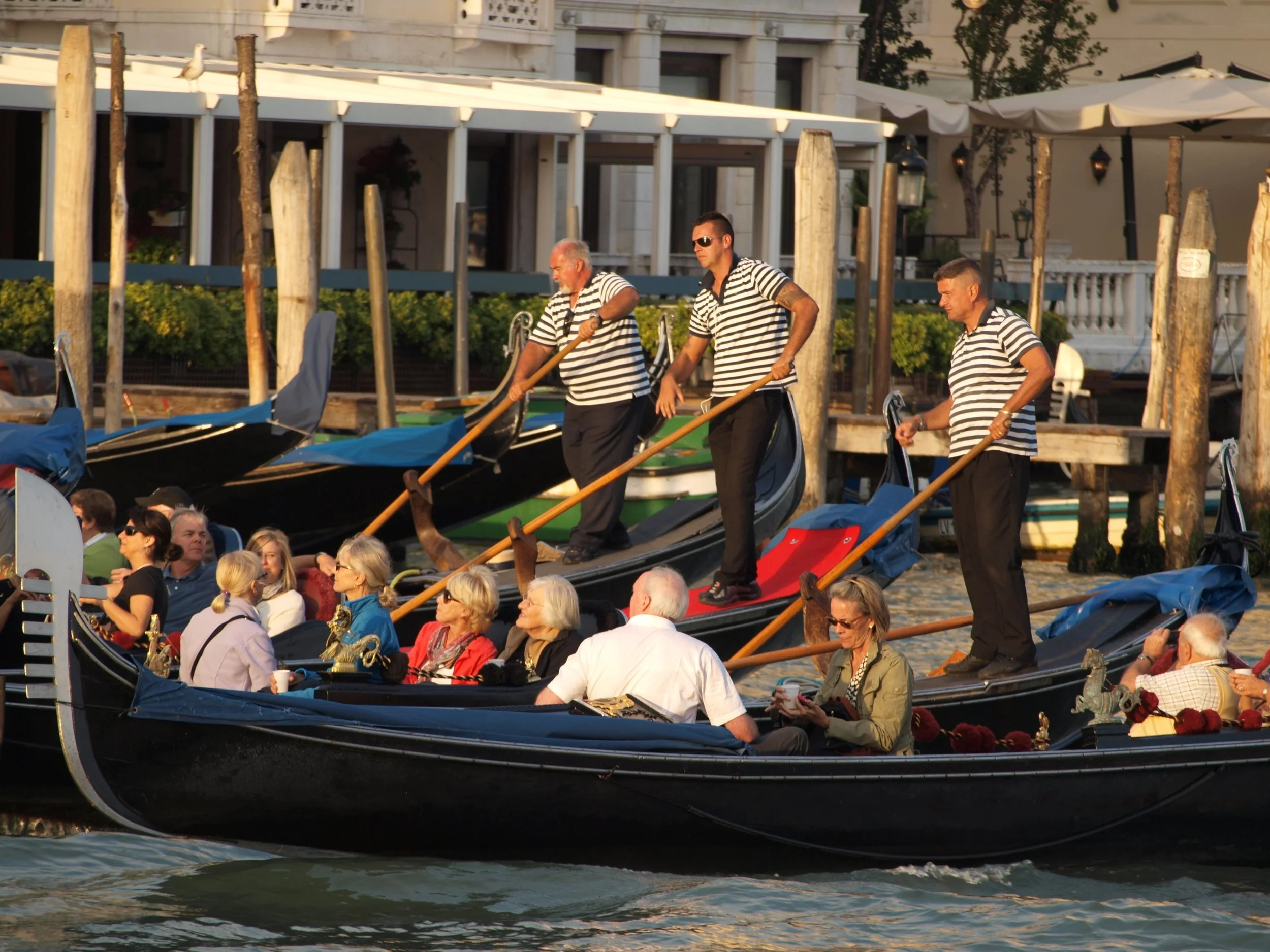 Gondoles à Venise naviguant sur un canal, avec des passagers et des gondoliers en uniformes rayés noirs et blancs, soleil couchant et bâtiments en arrière-plan.