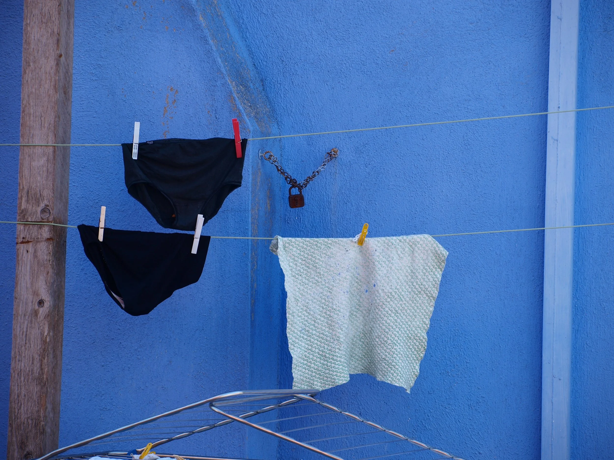 Ligne à linge avec vêtements suspendus, notamment un maillot de bain noir, un linge blanc avec motifs verts, une serviette ou une couverture moirée, fixés avec des pinces, devant un mur bleu.