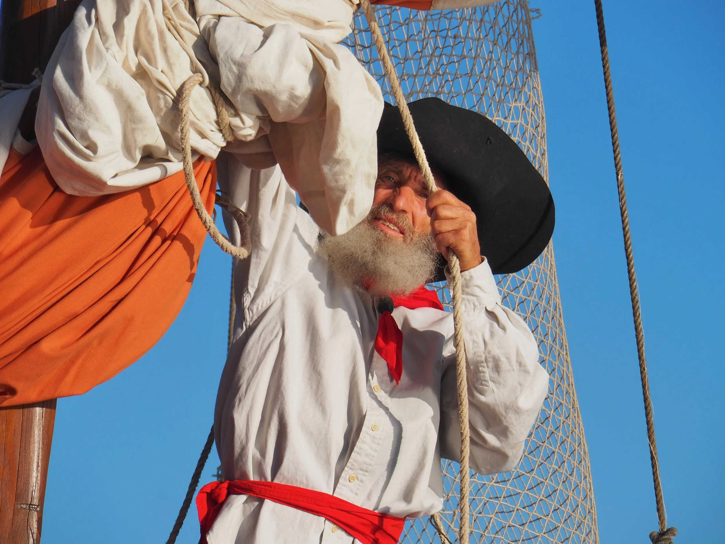 Un homme âgé, avec une barbe blanche, portant un grand chapeau noir, un uniforme blanc avec un foulard rouge, grimpe à une cordée sur une structure en treillis en métal, sous un ciel bleu clair.