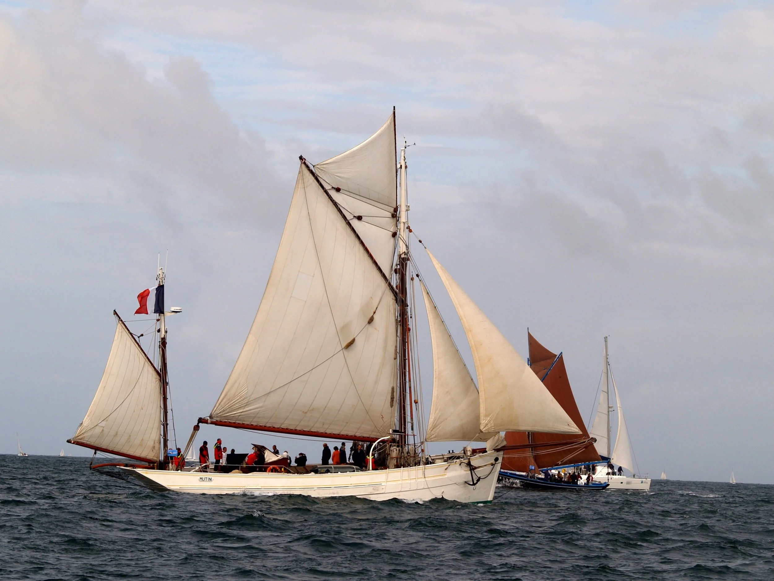 Voilier traditionnel avec plusieurs voiles en pleine mer, un groupe de personnes à bord, drapeau français en arrière-plan, ciel nuageux.