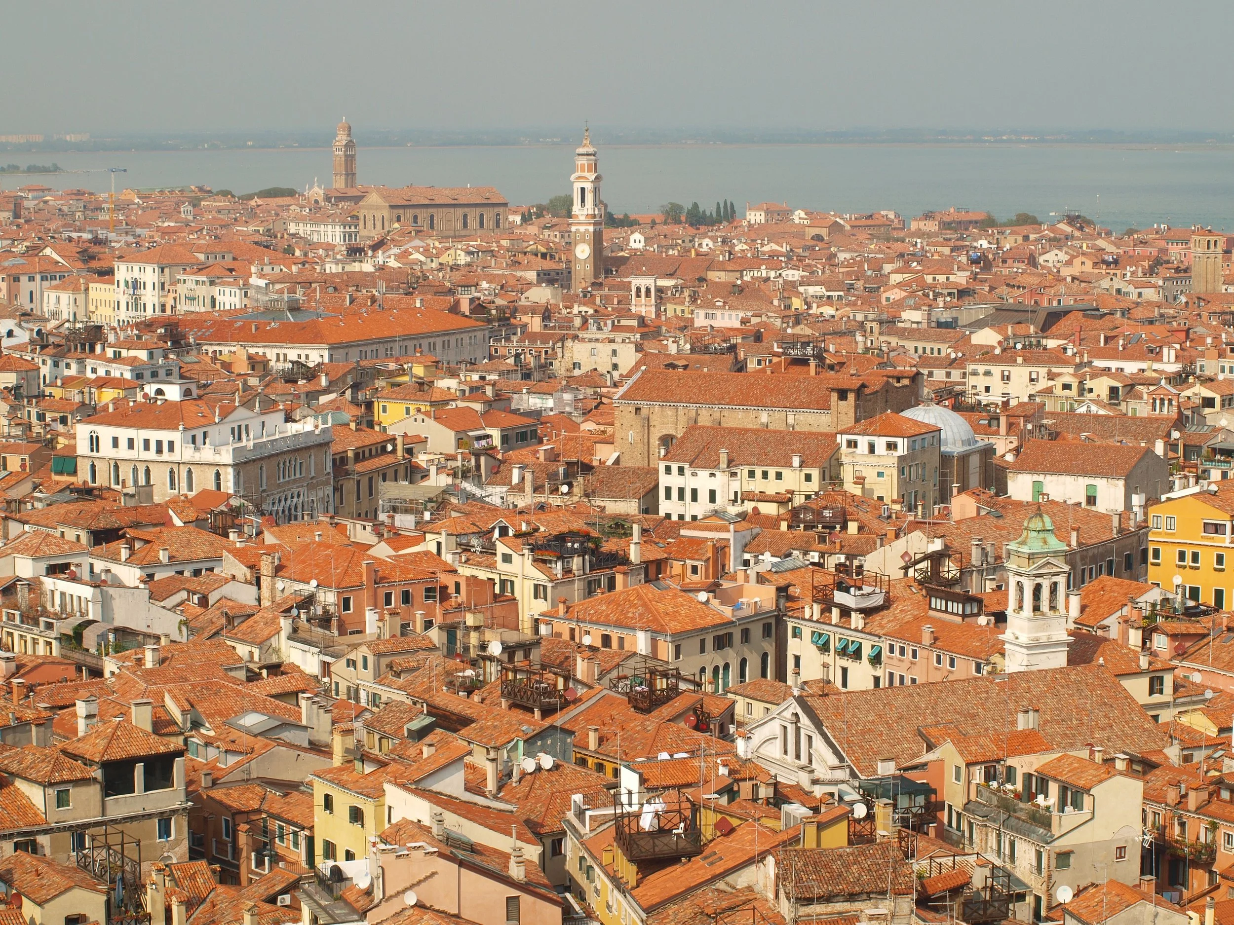 Vue panoramique sur un centre-ville européen avec de nombreux bâtiments aux toits en tuiles orange, incluant des clochers et des tours, en face d'un lac ou une mer calme, sous un ciel clair.