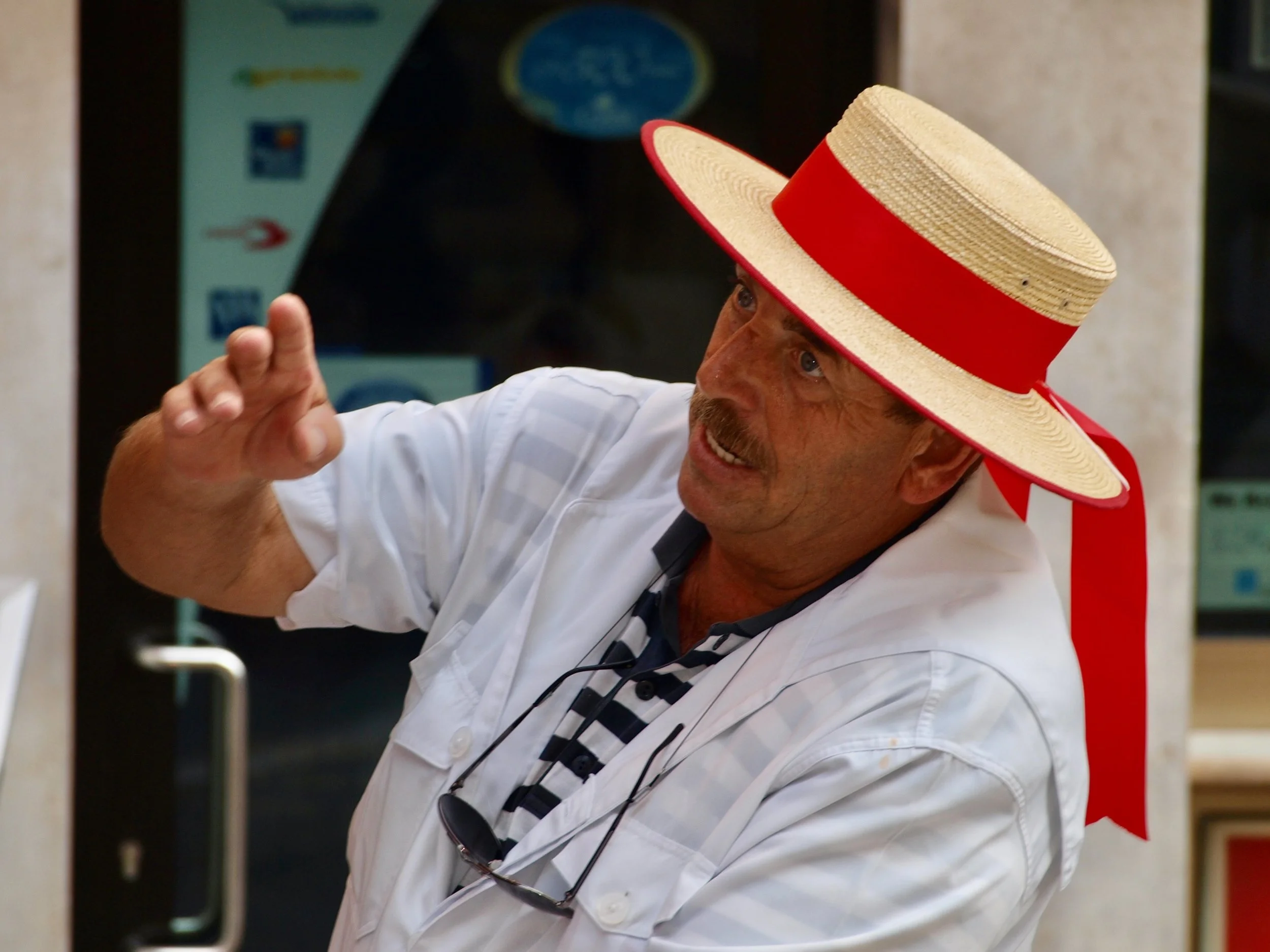 Un homme portant un chapeau de plage beige avec une bande rouge, un t-shirt blanc et des lunettes suspendues autour du cou, fait un geste de la main, probablement dans un environnement intérieur.