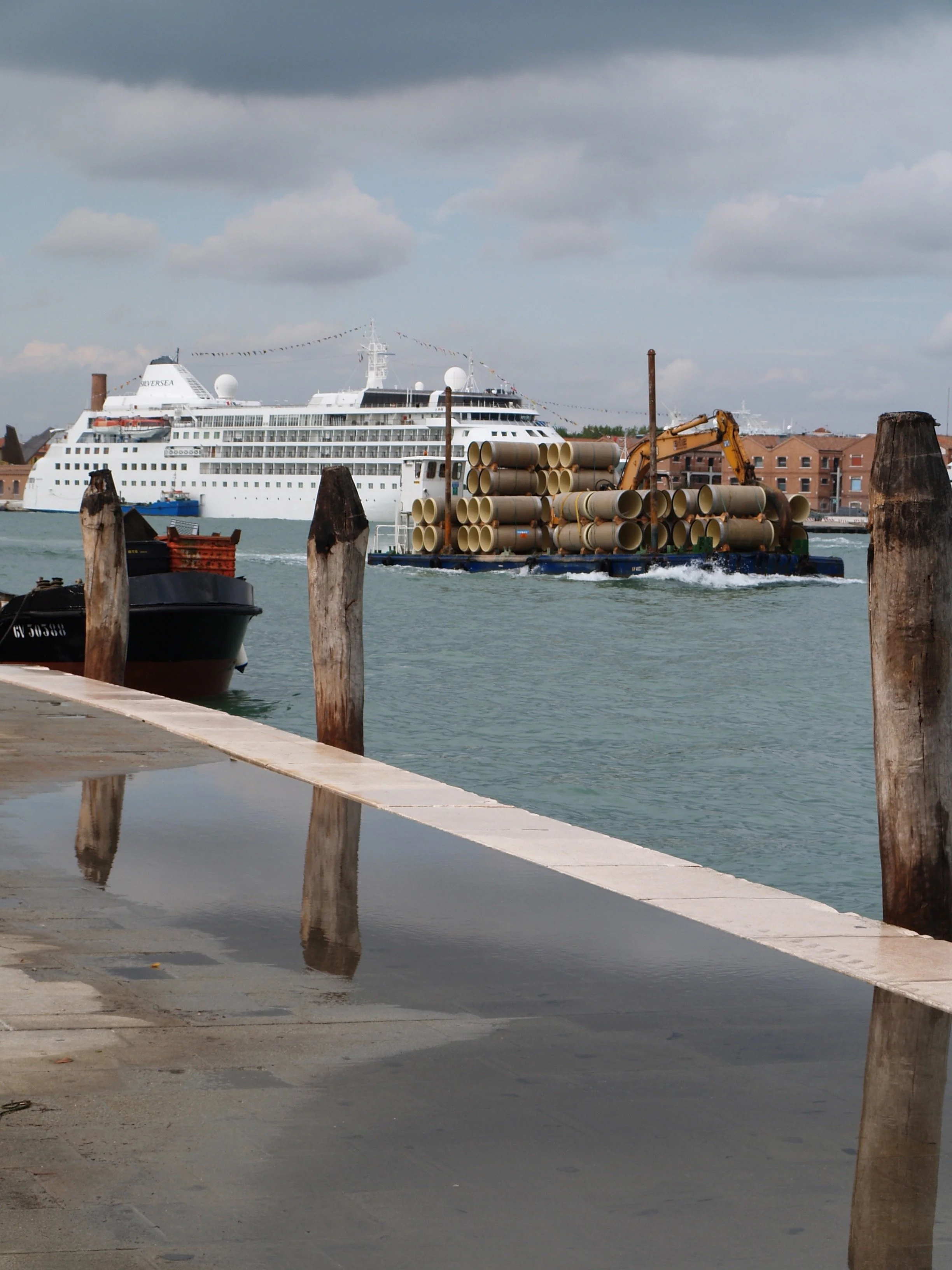 Un bateau de croisière blanc naviguant dans un port avec un ciel nuageux, au premier plan il y a des piquets en bois et des tuyaux de construction sur une barge.