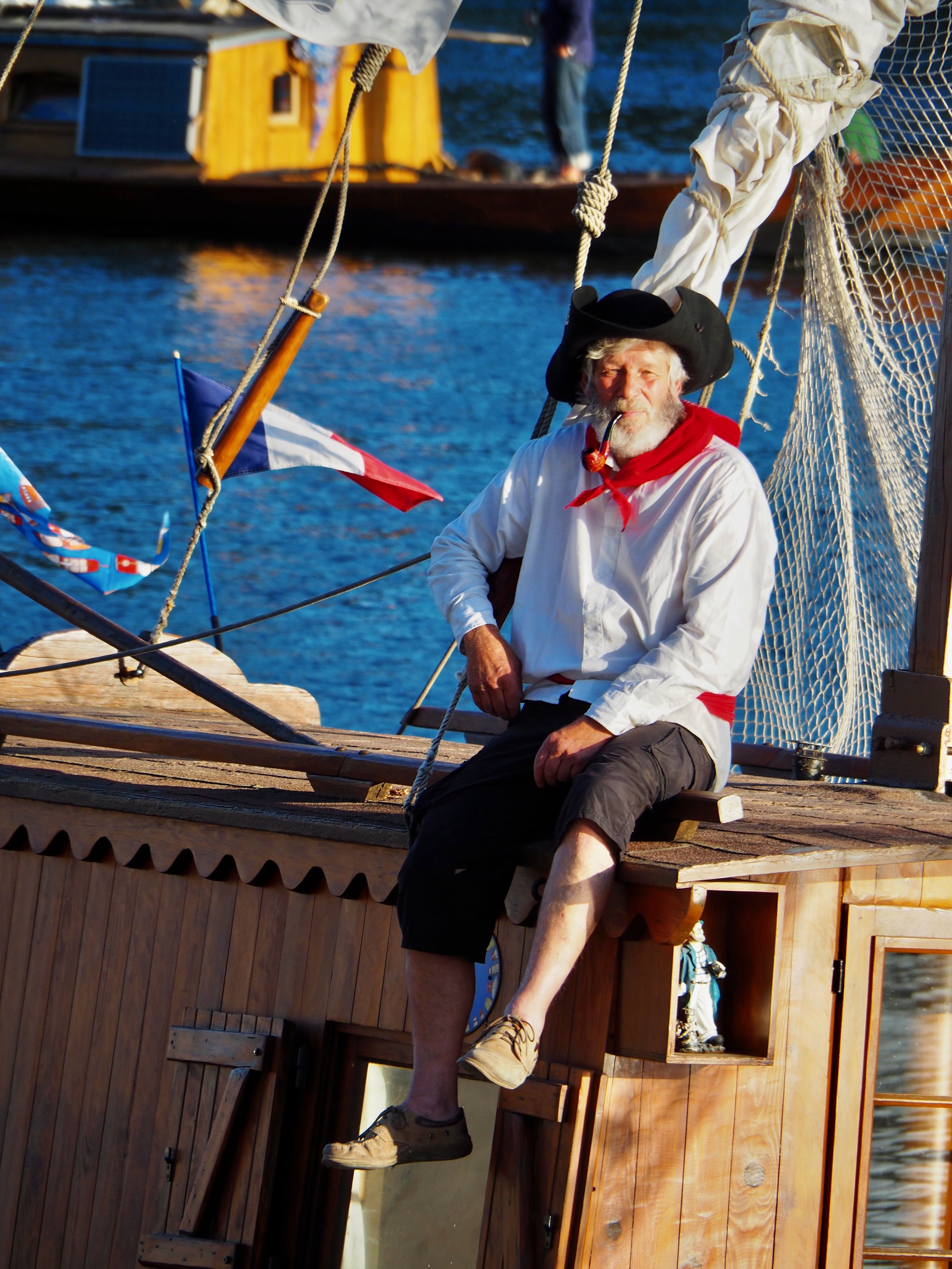 Un homme habillé comme un marin ou un pirate, avec un chapeau noir, une pipe, un chemise blanche, un foulard rouge, et un short noir, assis sur le pont d'un bateau en bois, avec la mer en arrière-plan.