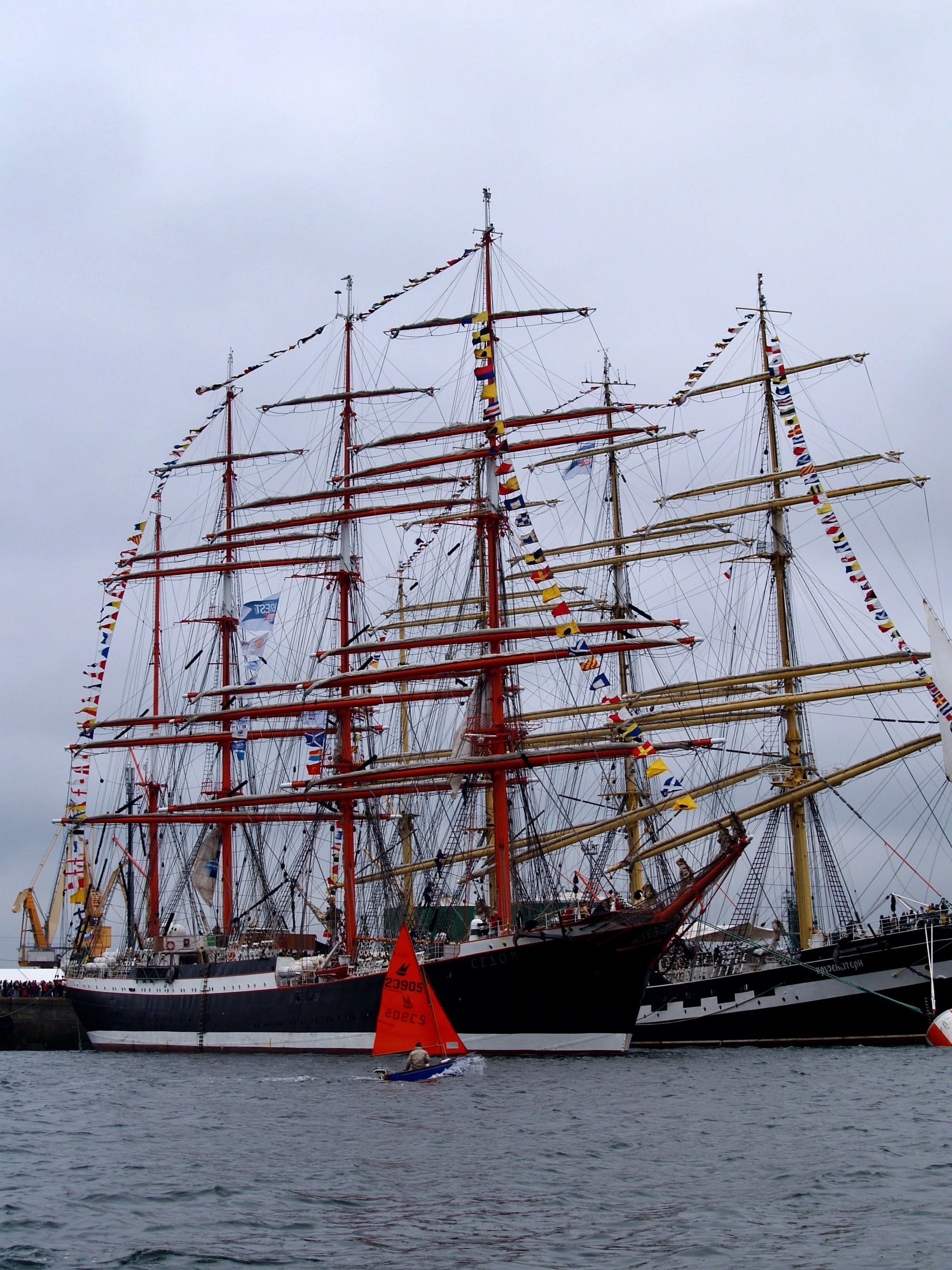 Une grande voile de navire à trois mats, décorée de fanions colorés, échoué sur un port avec une personne dans un petit bateau orange à l'avant, sous un ciel nuageux.