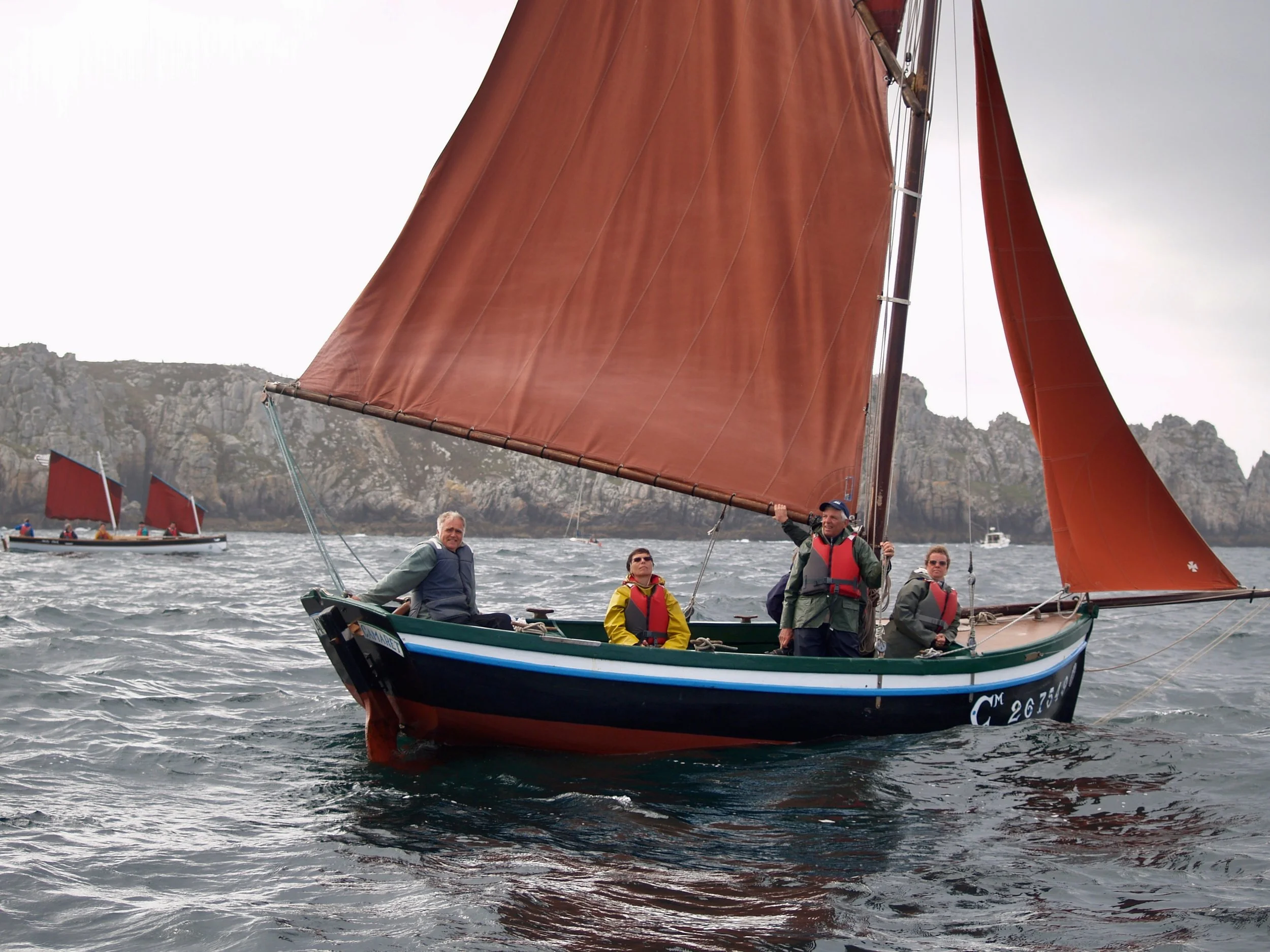 Groupe de personnes sur un voilier avec voiles orange, en mer avec des falaises en arrière-plan.