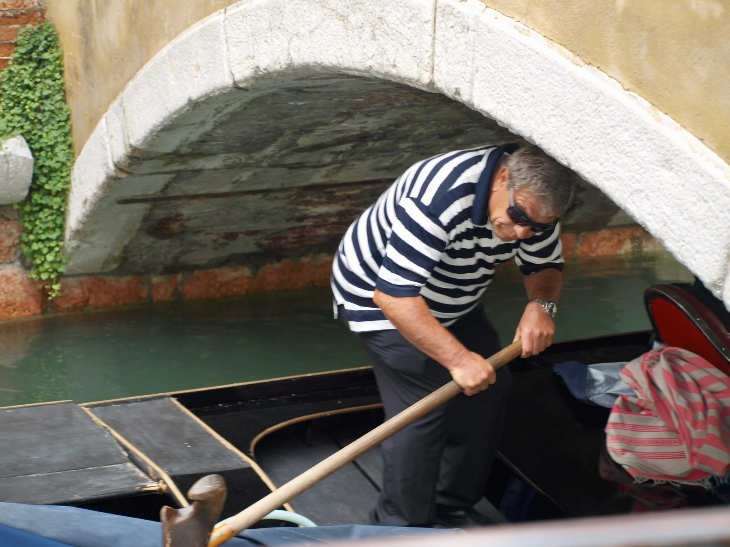 Une personne portant une chemise à rayures noires et blanches utilise un bâton pour pousser une personne assise dans un bateau en bois. La scène est à l'intérieur d'un bâtiment ou d'une structure en pierre avec une arche visible au plafond.