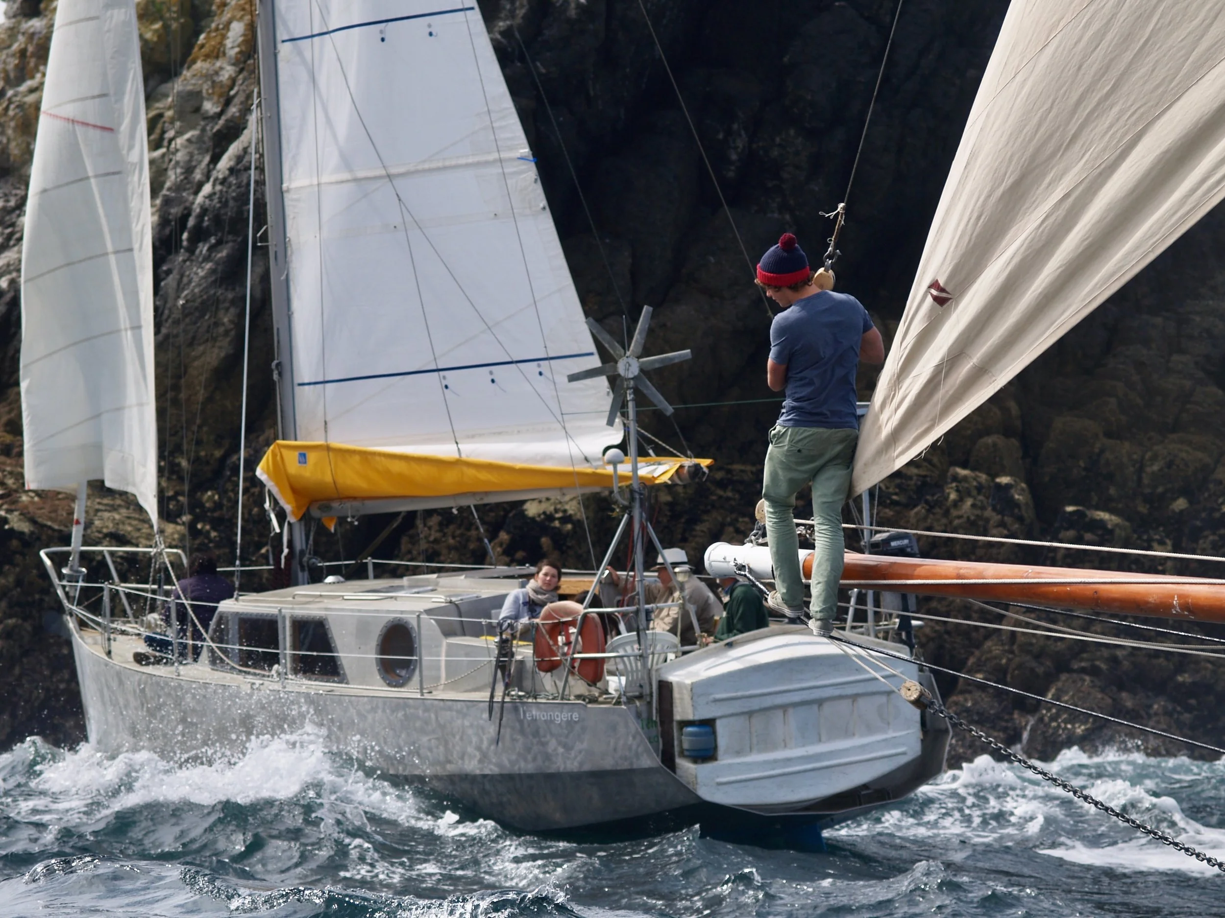 Un voilier blanc naviguant en mer avec un homme en t-shirt bleu et bonnet rouge, des autres voiliers en arrière-plan et une falaise rocheuse.