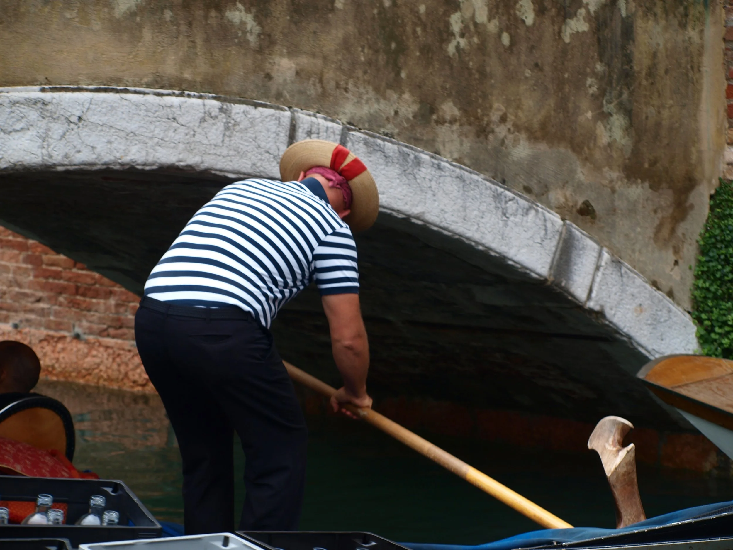 Personnage en costume de marin avec un chapeau en paille portant un râteau dans un canal, vue partielle de son dos, entouré de végétation et de bâtiments anciens.