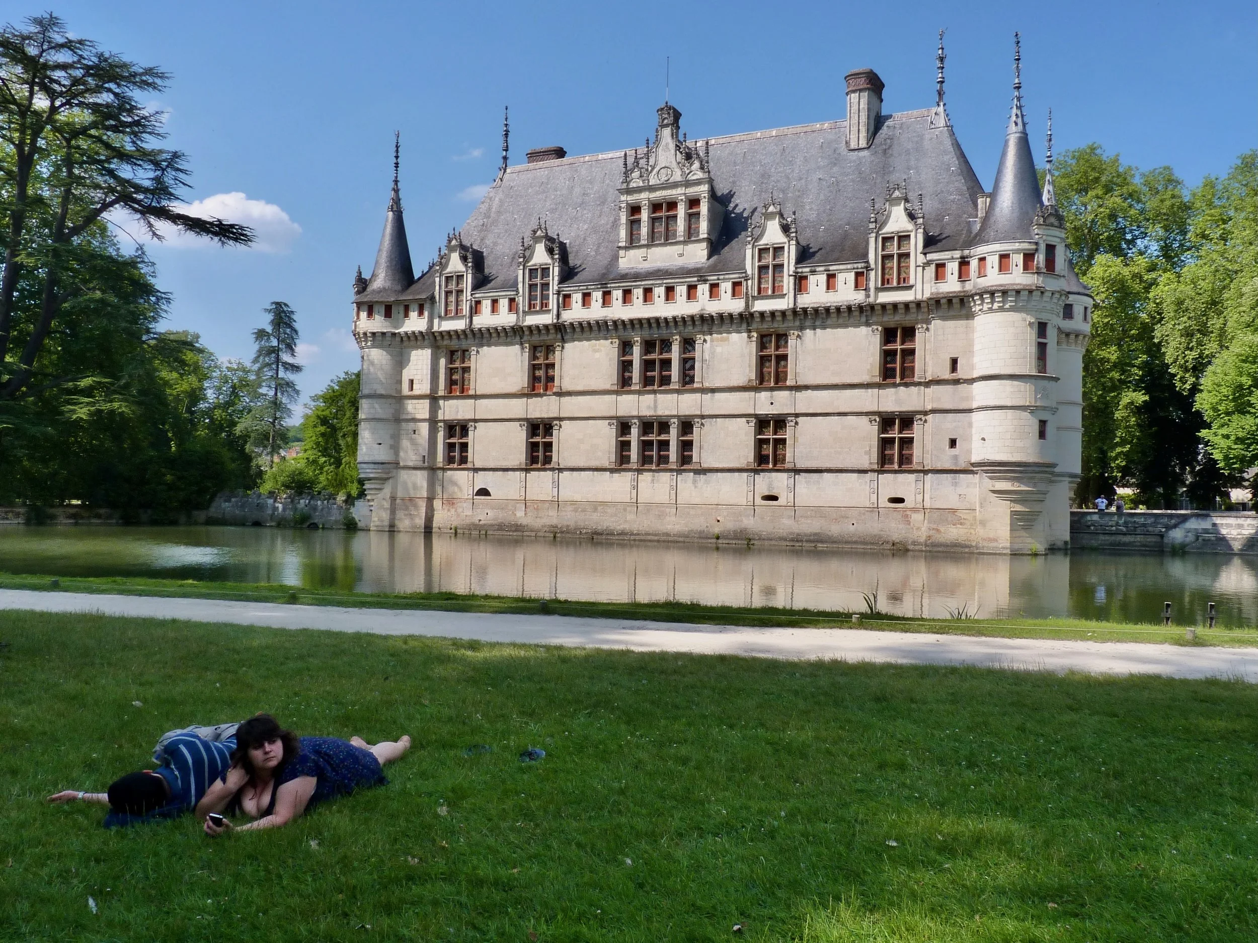 Une étudiante allongée sur l'herbe dans un parc, avec un château en pierre visible en arrière-plan, entouré d'arbres et d'une rivière.