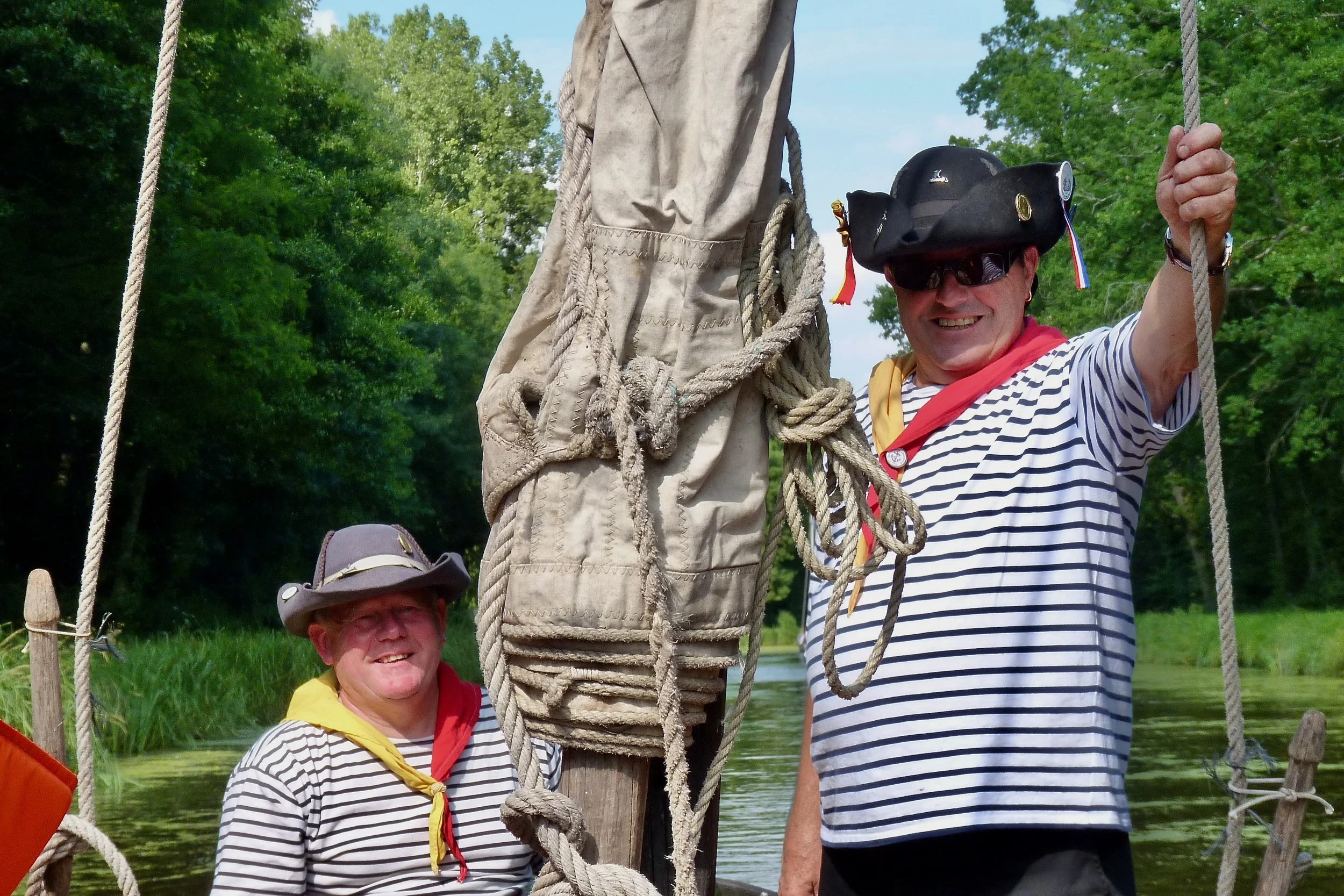 Deux hommes habillés en marins rayés posent sur une barque en bois sous un arbre vert. L'un porte un chapeau en paille et un foulard jaune, l'autre porte un chapeau noir avec des médailles et des lunettes de soleil.