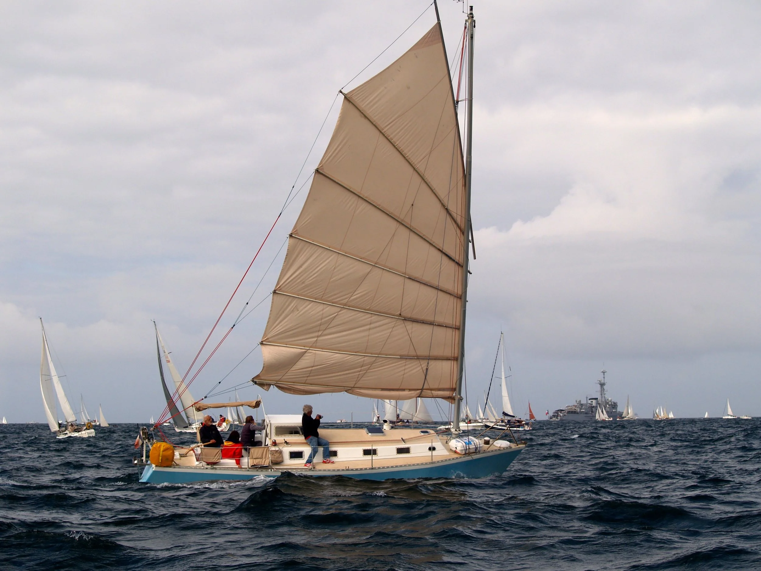 Un voilier avec une voile beige naviguant en mer lors d'une régate, avec plusieurs autres voiliers en arrière-plan et un grand navire. Le ciel est nuageux.
