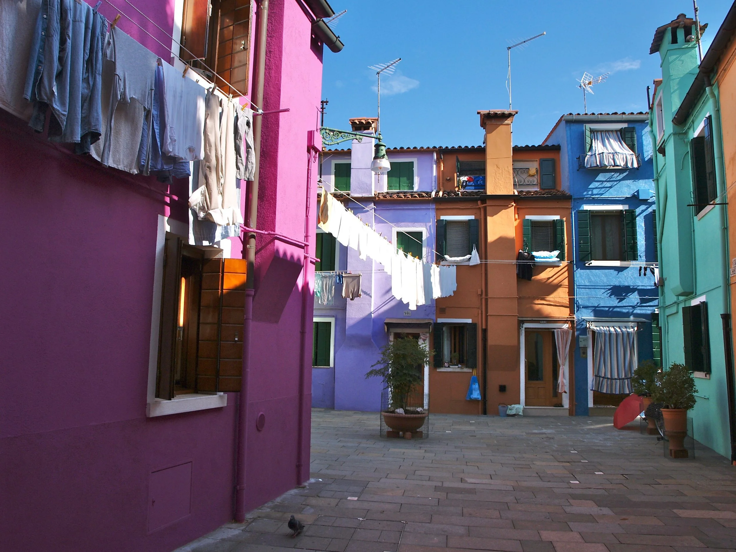 Cour intérieure avec bâtiments colorés en orange, violet, bleu et vert, linge suspendu pour sécher, volets verts, parasol, plantes en pot et ciel bleu en arrière-plan.