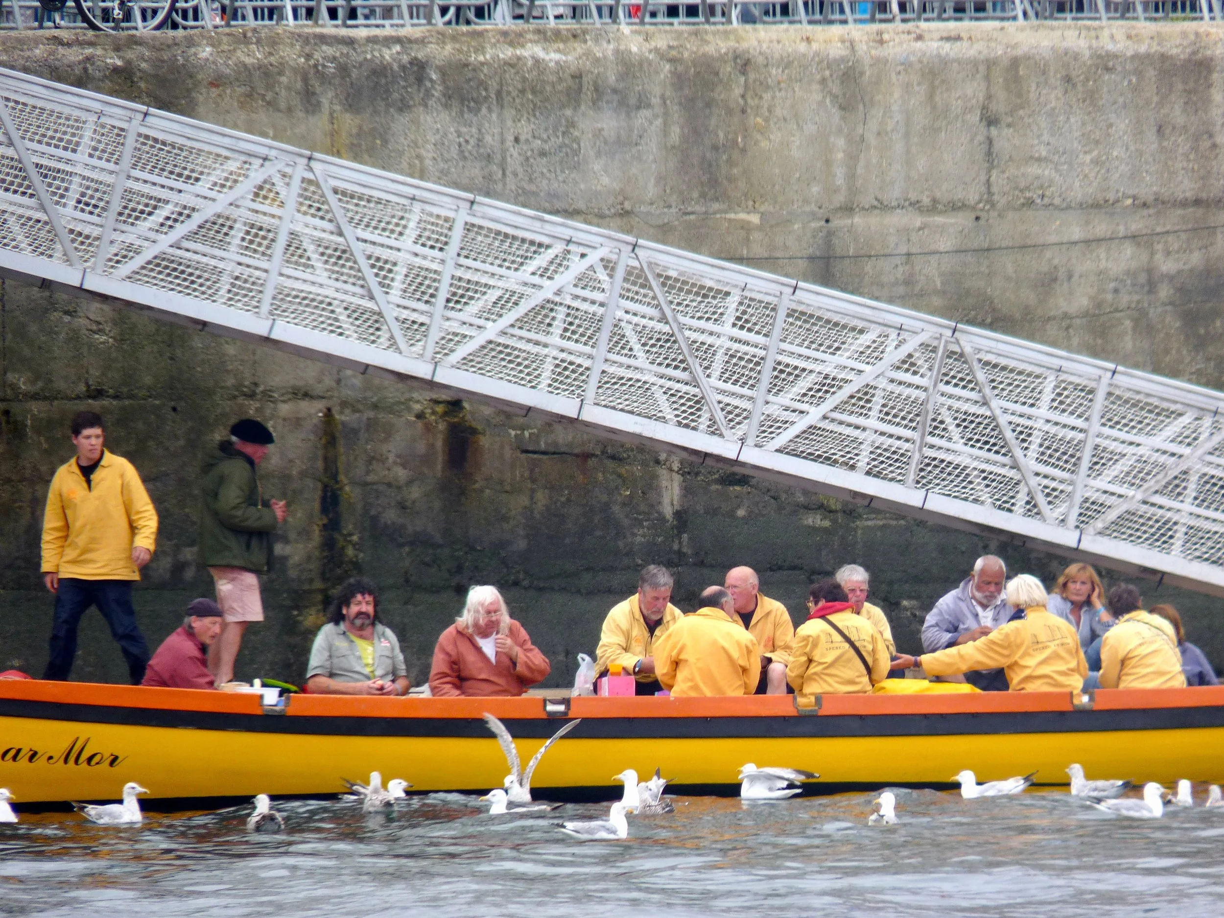 Groupe de personnes portant des gilets jaunes et des vêtements variés, assises ou se tenant près d'un bateau sur l'eau, avec une falaise en arrière-plan et des mouettes flottant à la surface de l'eau.