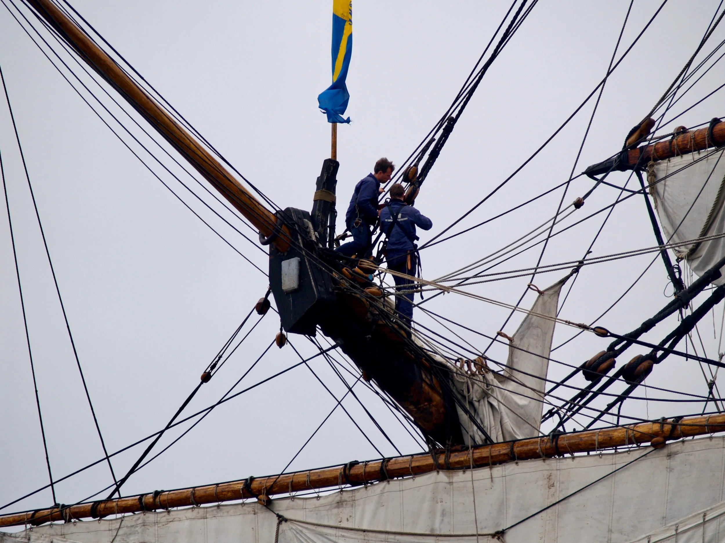 Deux hommes travaillent sur la mâture d’un vieux navire à voiles, entourés par un réseau complexe de cordages.
