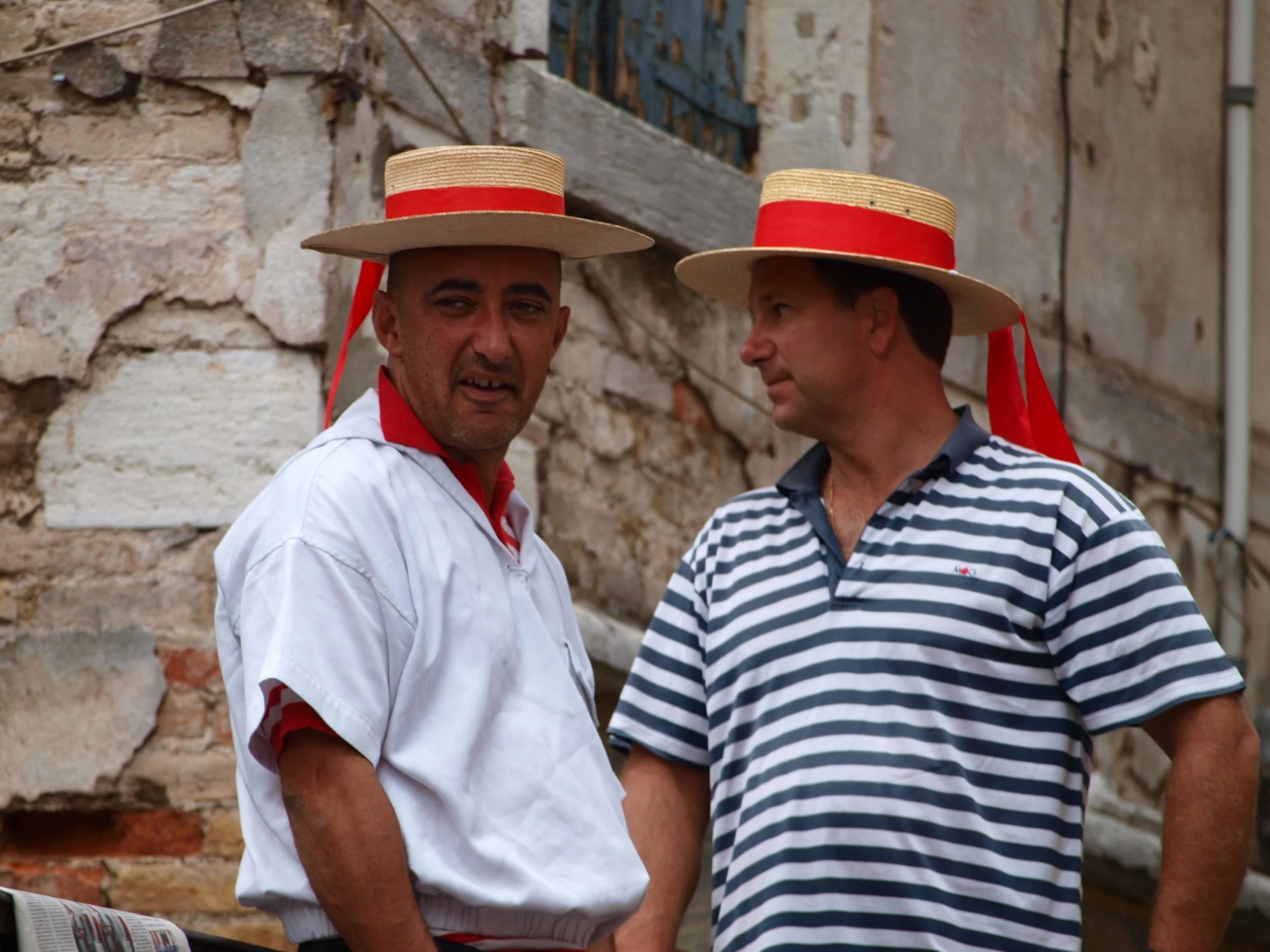 Deux hommes portant des chapeaux de style canotier avec un ruban rouge, se tenant devant un mur en pierre usée, en conversation ou en regardant quelque chose.