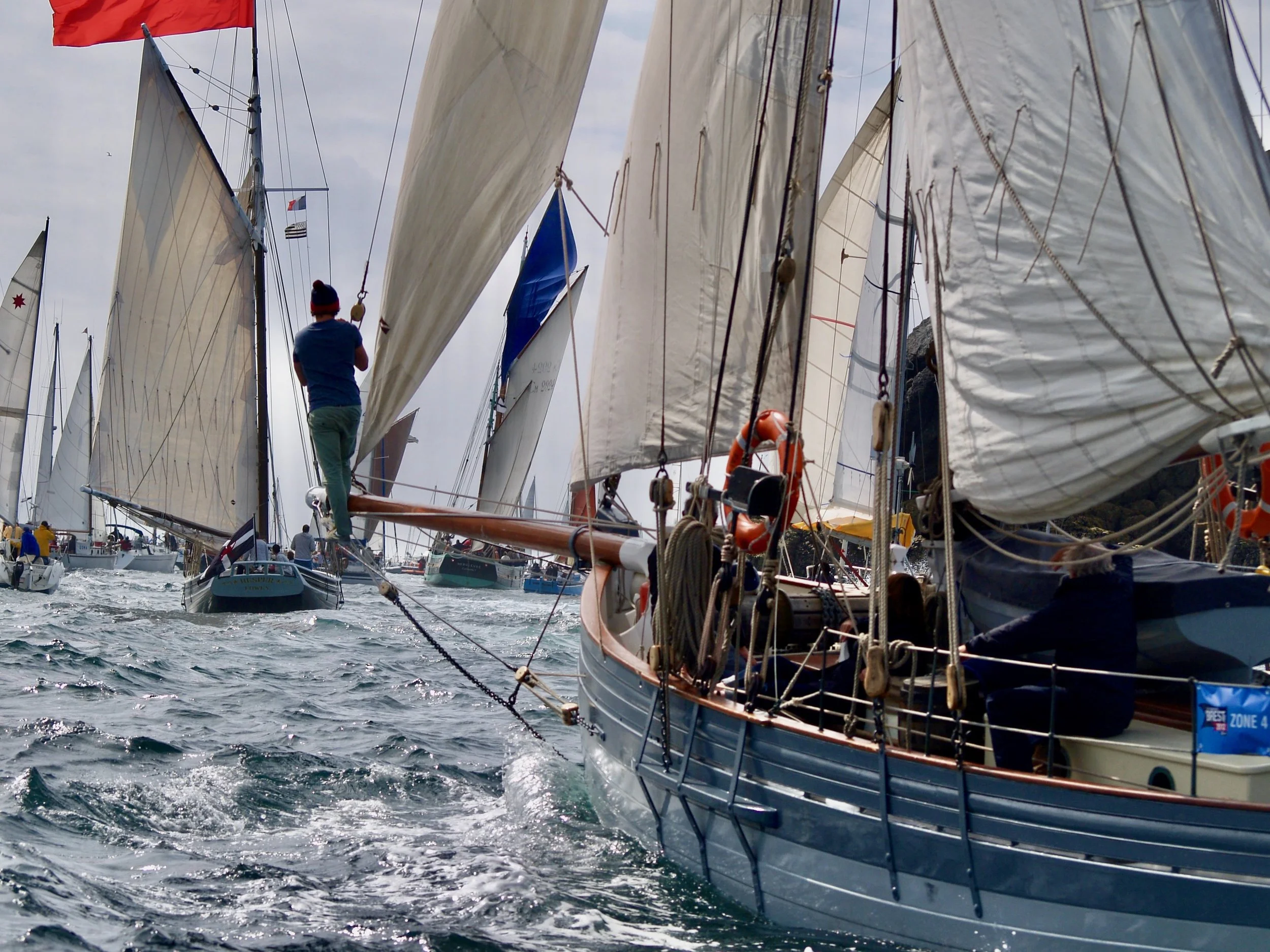Bateaux à voile alignés lors d'une régate, vue entre plusieurs voiles avec un homme en haut d'un bateau, portant un bonnet, observant l'action.