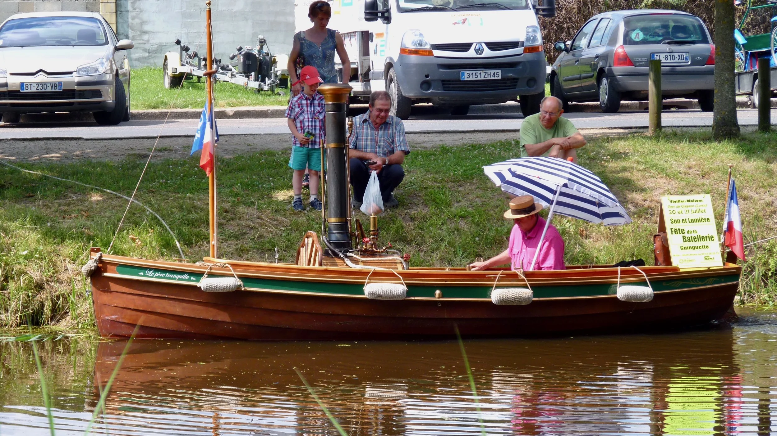 Une scène de fête avec un bateau en bois dans l'eau, des personnes assises sur le rivage et un stand avec une affiche promotionnelle à côté.