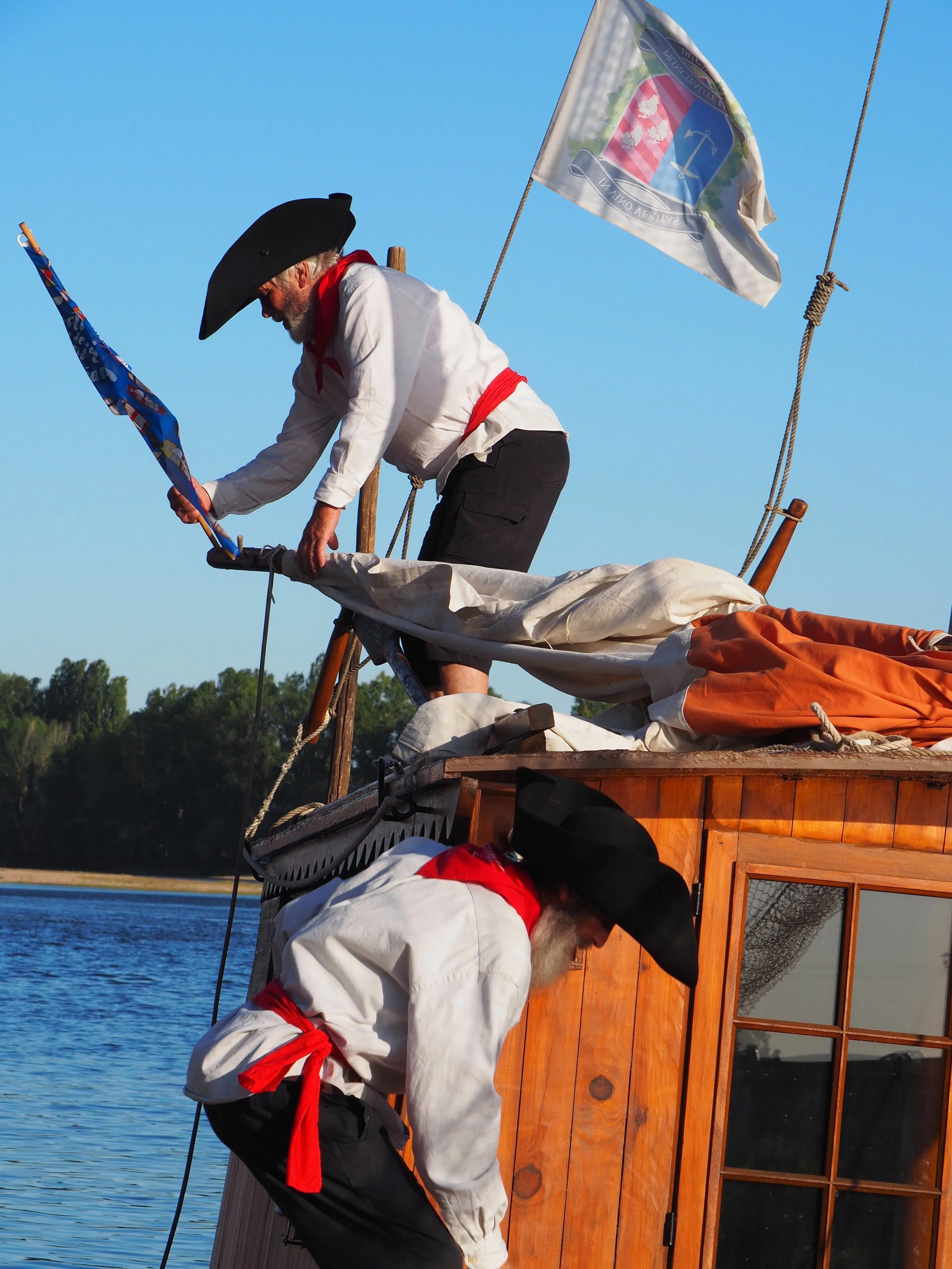 Trois hommes habillés en costumes traditionnels avec des chapeaux noirs et des foulards rouges, chargés de drapeaux, sur un bateau en bois sur l'eau, en train de préparer quelque chose.