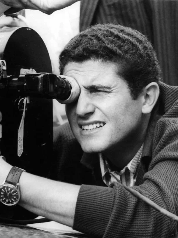 Un homme regarde à travers un télescope, en utilisant son œil droit, avec une expression concentrée et souriante.