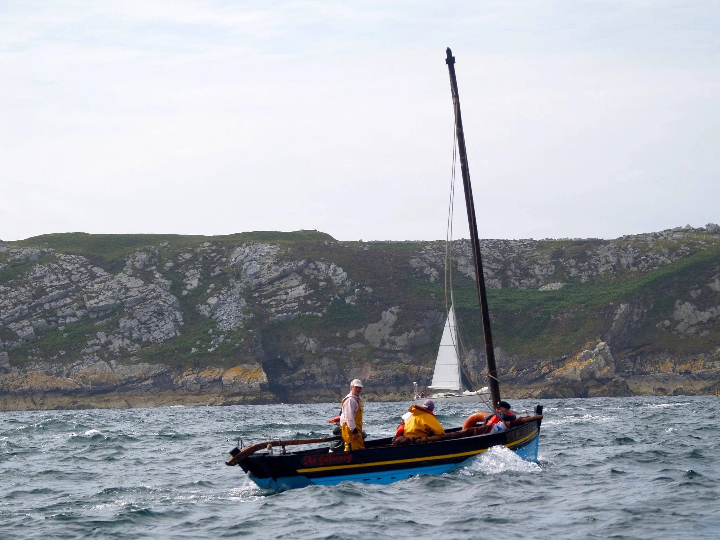 Une embarcation avec plusieurs personnes portant des vêtements de sécurité, naviguant en mer près d'une côte rocheuse et verdoyante, avec un homme en avant du bateau et un voilier en arrière-plan.