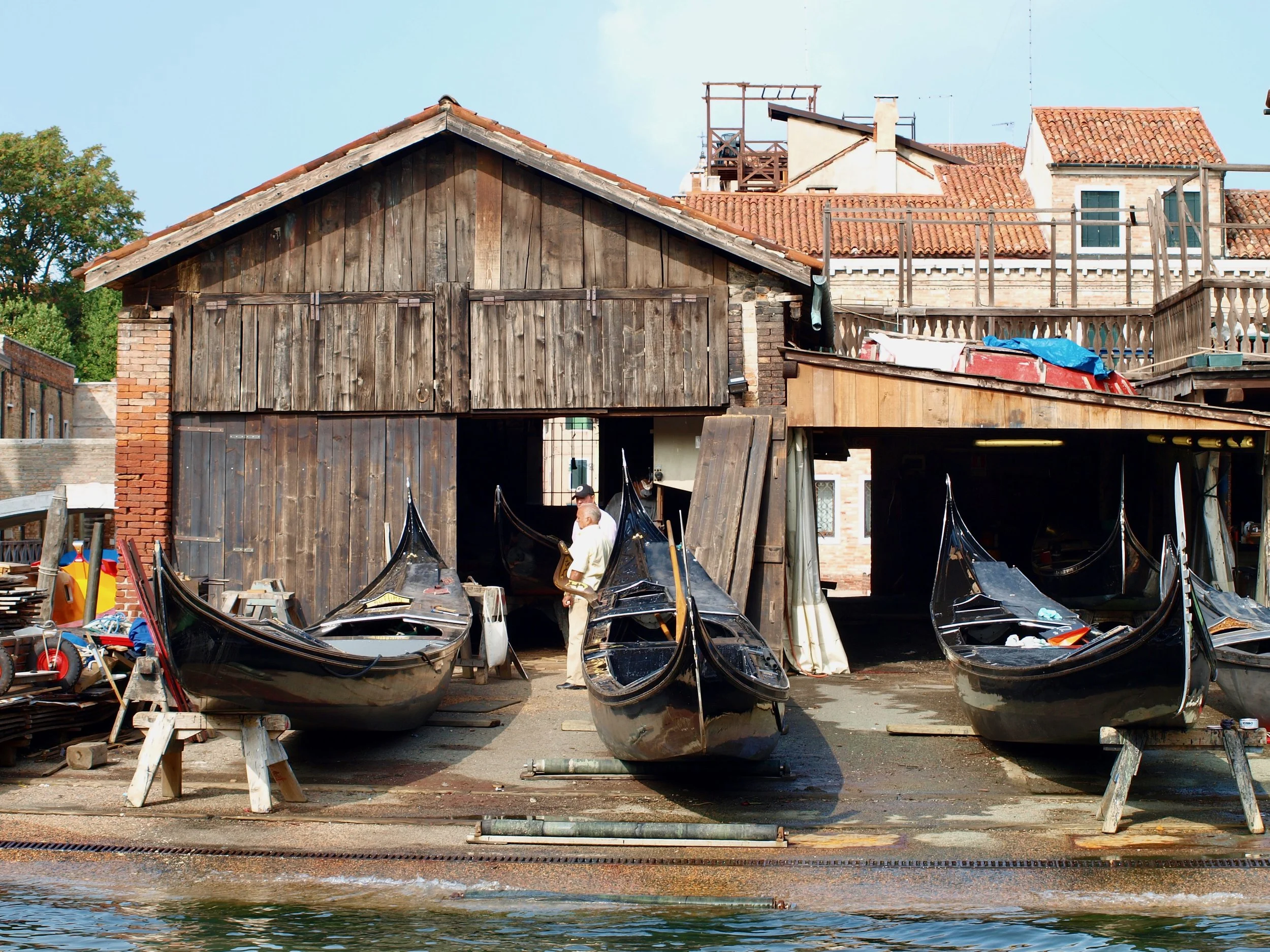 Un quai avec des bateaux noirs amarrés, derrière un bâtiment en bois et en briques sous un ciel bleu. On voit des maisons avec des toits en tuiles rouges au fond.
