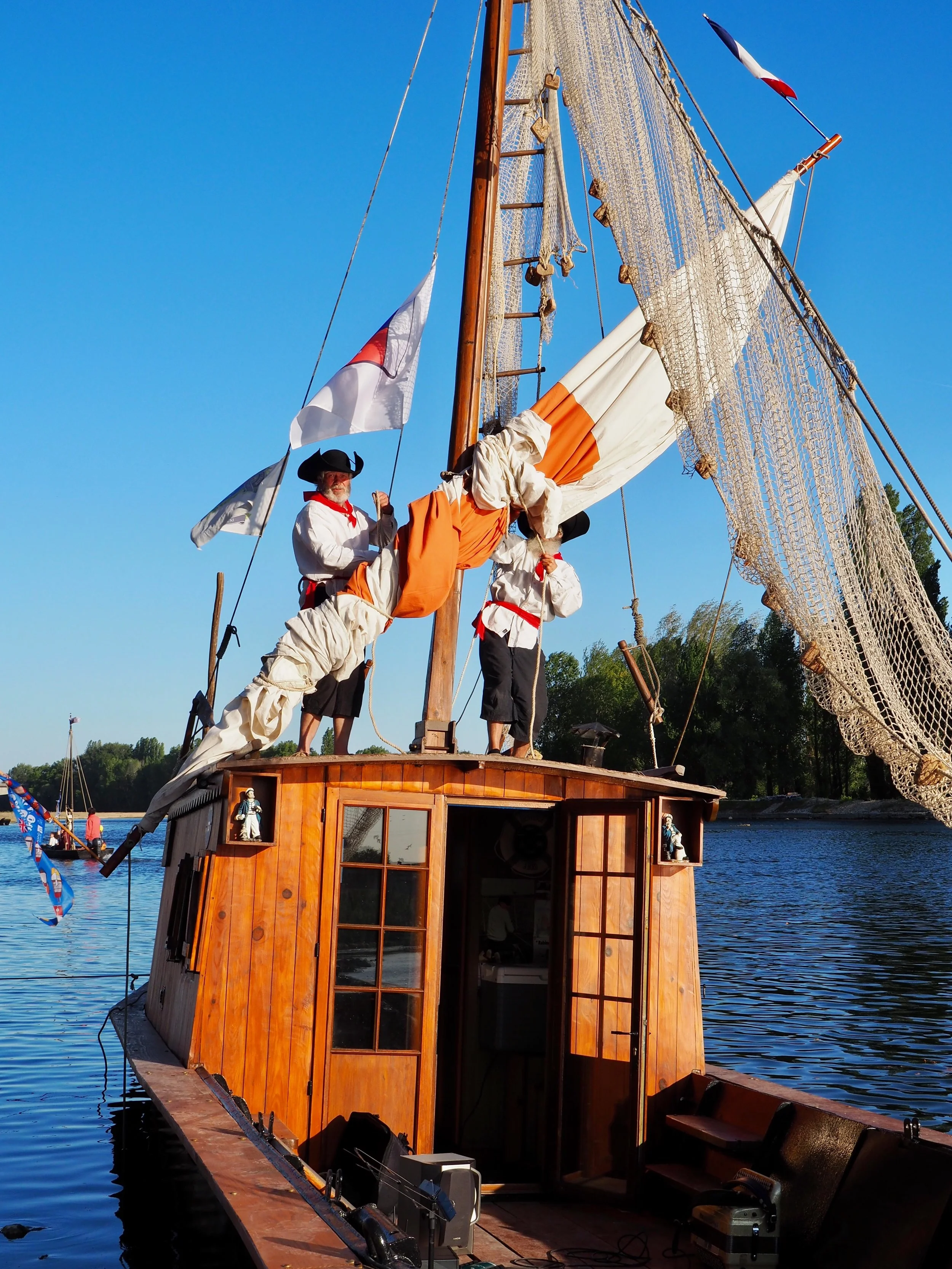 Un navire en bois avec des voiles et des matelots en costume historique sur un fleuve, avec un ciel clair et des arbres en arrière-plan.