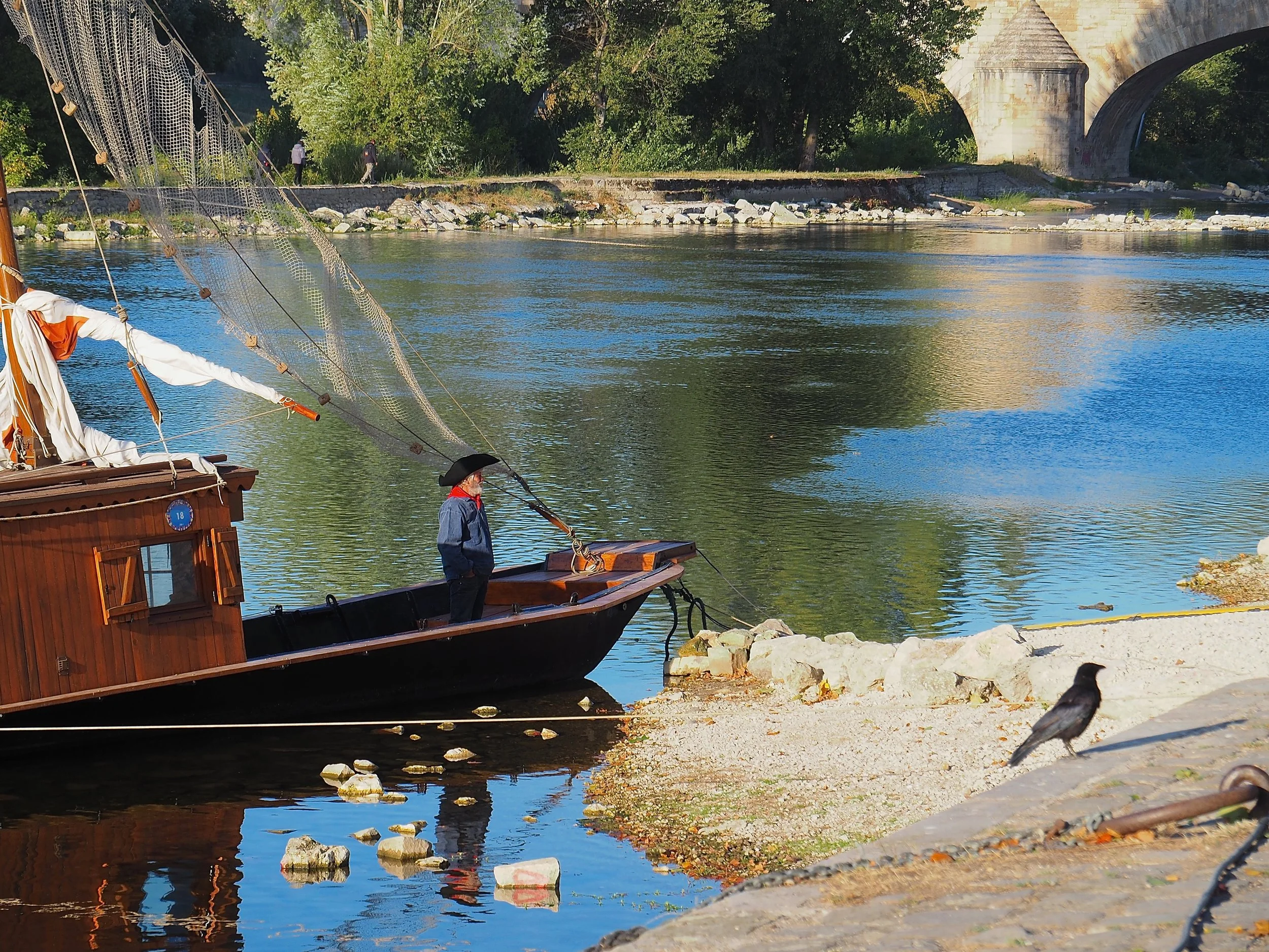Un bateau en bois amarré au bord de la rivière avec un homme en costume et chapeau, des passants se promènent sous un pont en pierre en arrière-plan, entouré d'arbres verts.