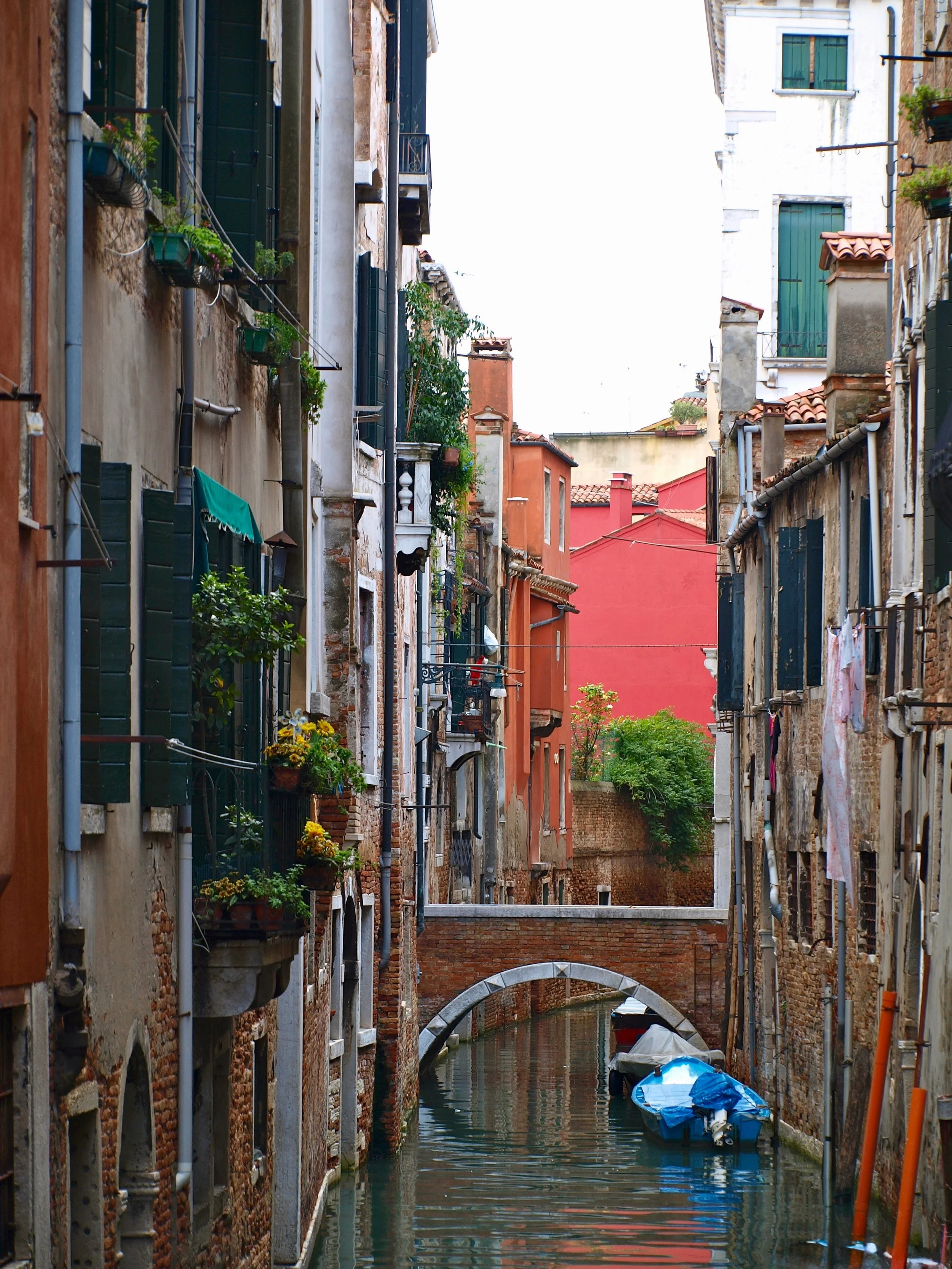 Une petite canalisation bordée de bâtiments anciens avec fenêtres à volets verts, quelques bateaux amarrés, un petit pont en pierre et des murs en briques et en stucco.