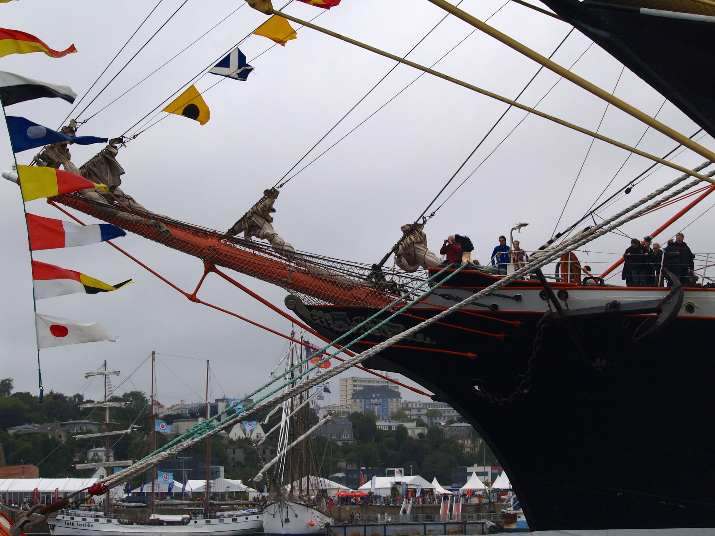 Vue d'un vieux voilier avec ses voiles et ses cordages, plusieurs personnes à bord et sur le quai, des tentes et drapeaux colorés lors d'un festival maritime.