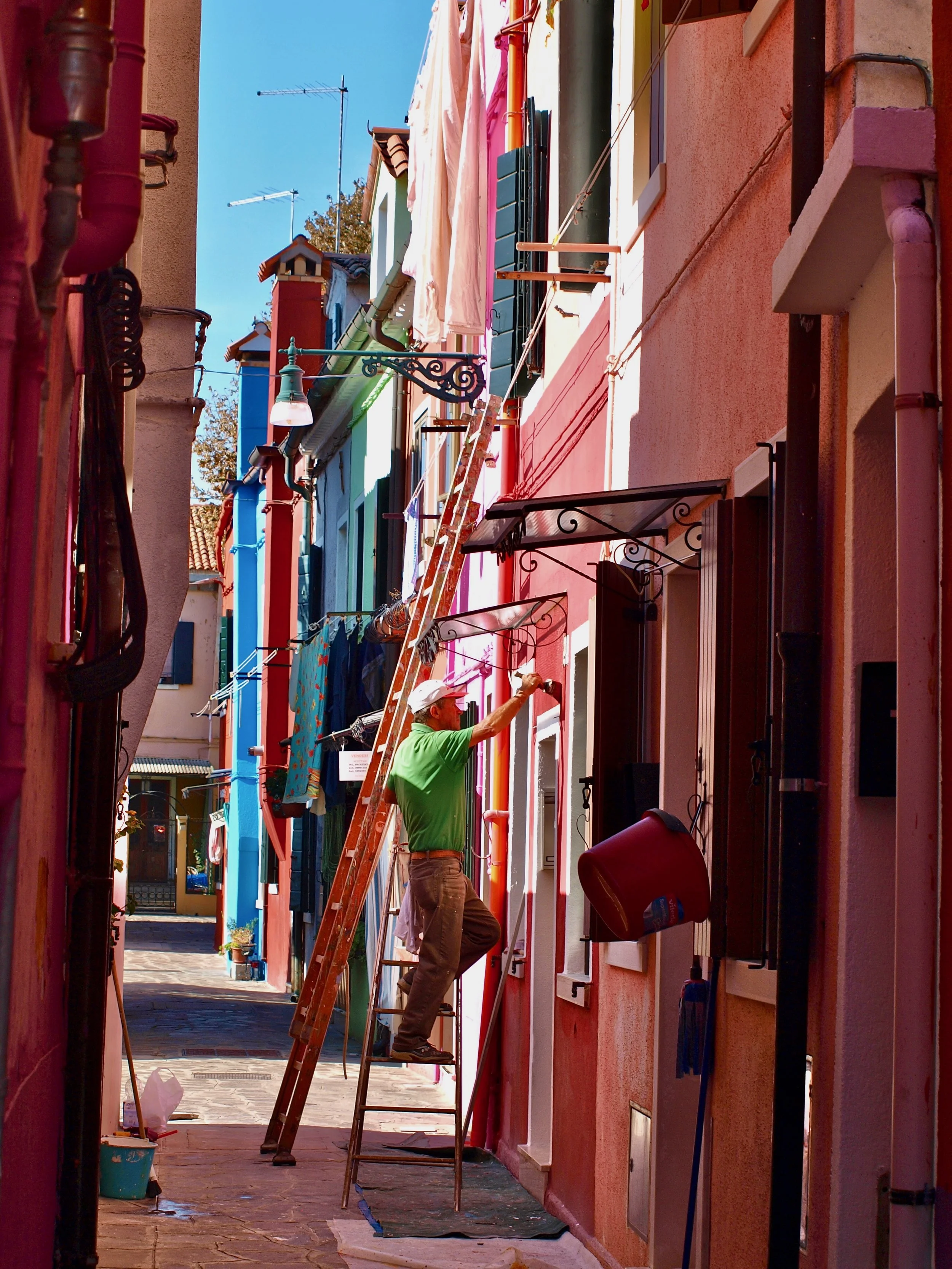 Un homme en vert peint la façade d'un bâtiment coloré en rose, utilisant une échelle et un pinceau, dans une rue étroite ensoleillée.