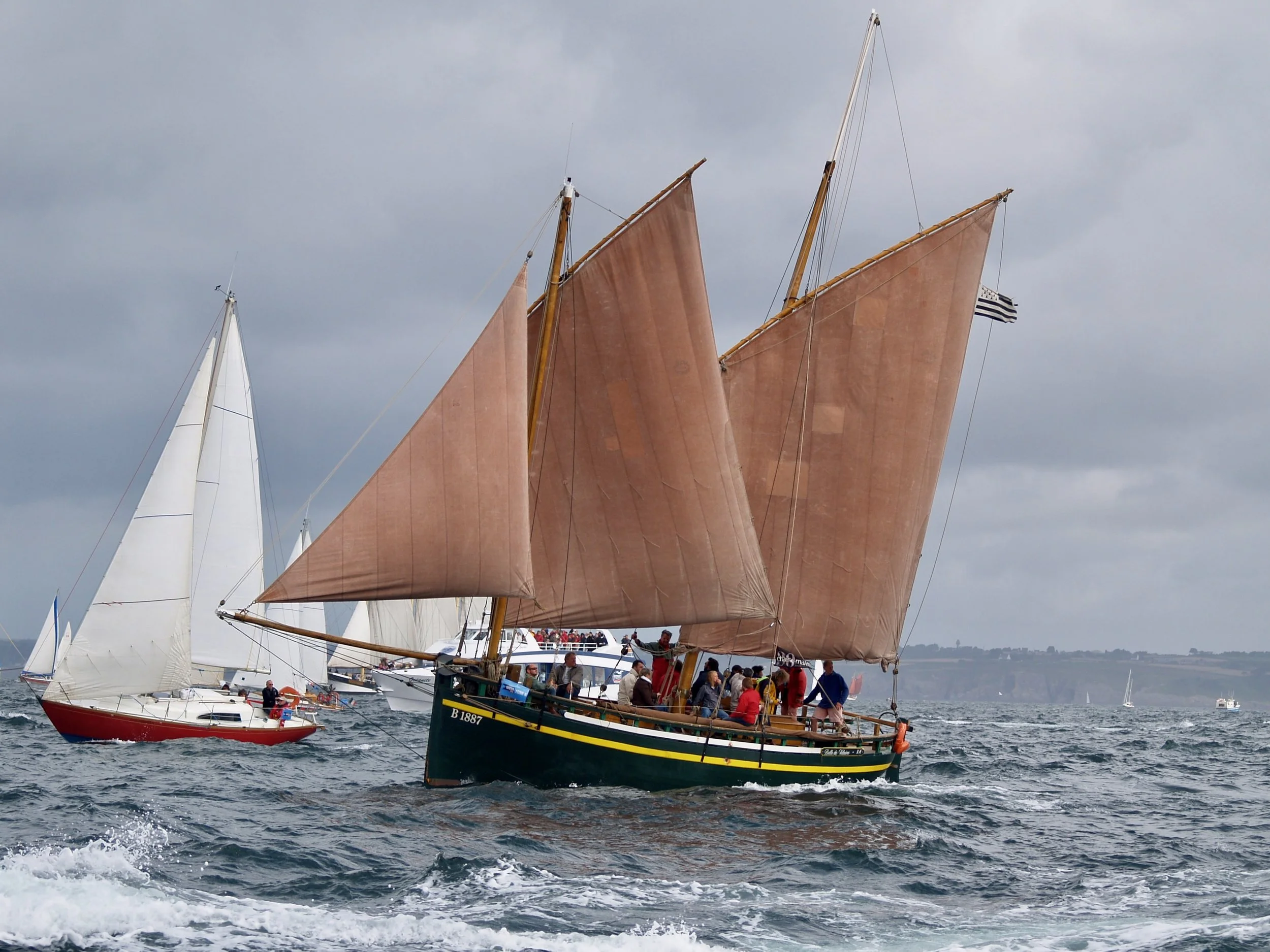 Un bateau traditionnel à voiles colorées navigue sur la mer avec plusieurs autres voiliers en arrière-plan. Des passagers sont visibles sur le bateau, et le ciel est nuageux.