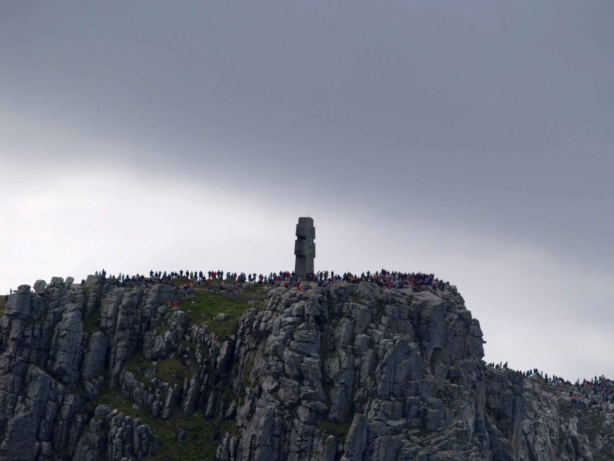 Une photographie d'un sommet de montagne avec un grand nombre de touristes rassemblés, sous un ciel nuageux et une tour de télécommunication en métal en arrière-plan.