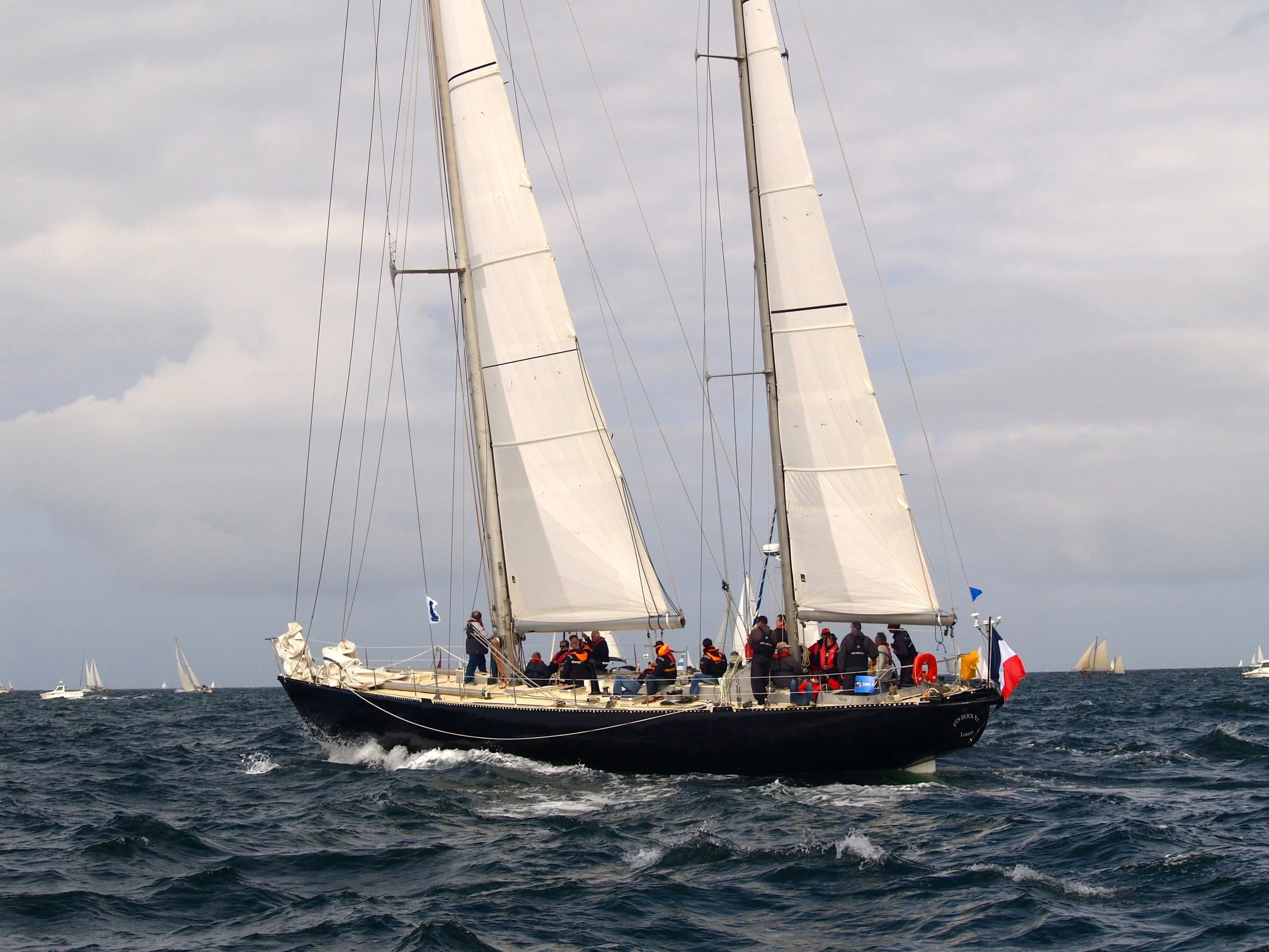 Un voilier naviguant en mer avec de nombreuses personnes à bord, portant des vestes de vie, et d'autres voiliers en arrière-plan sous un ciel nuageux.