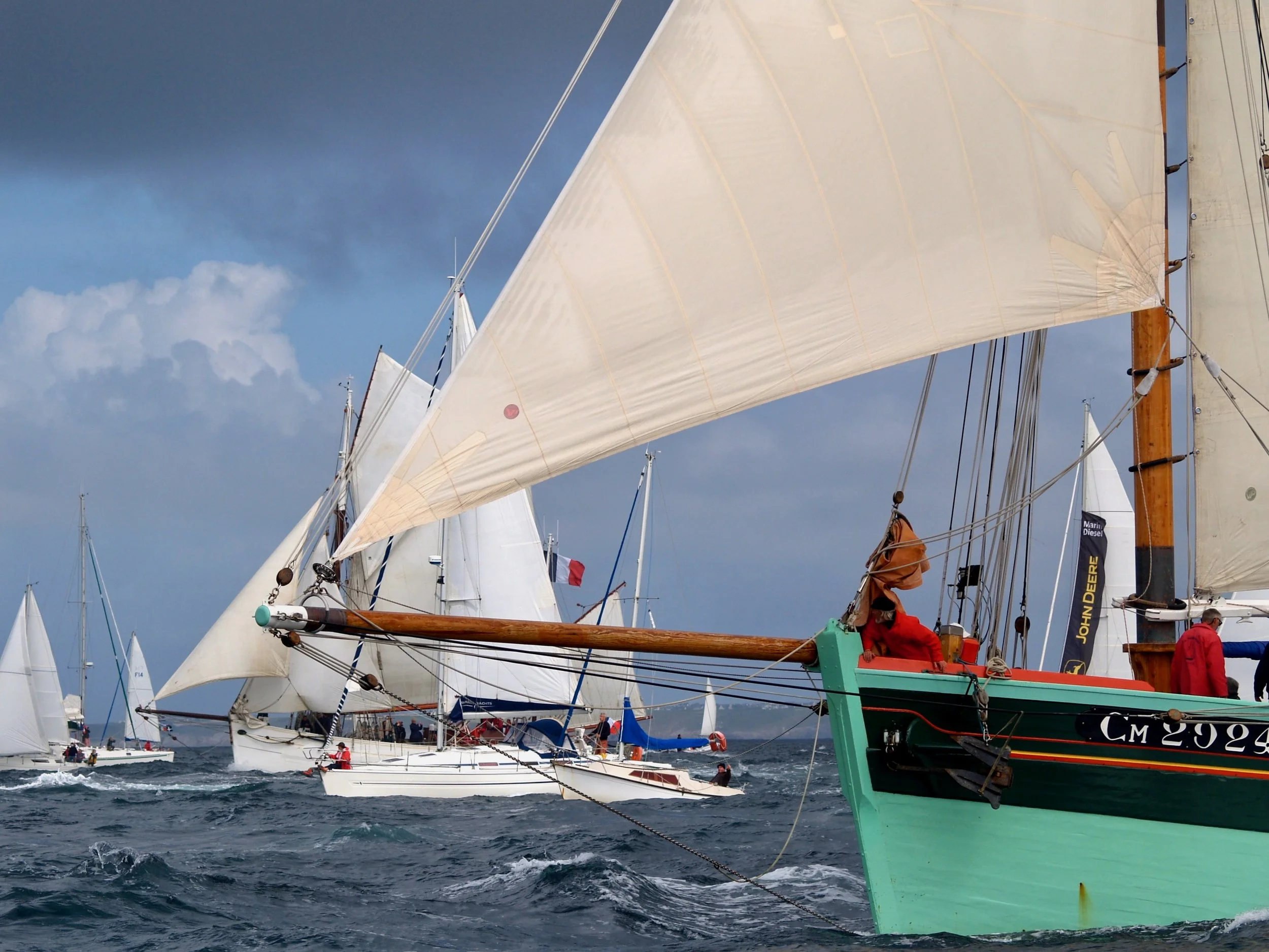 Des voiliers naviguant en mer avec leurs voiles déployées, certains avec des mâts en bois. Le ciel est partiellement nuageux et la mer est agitée.