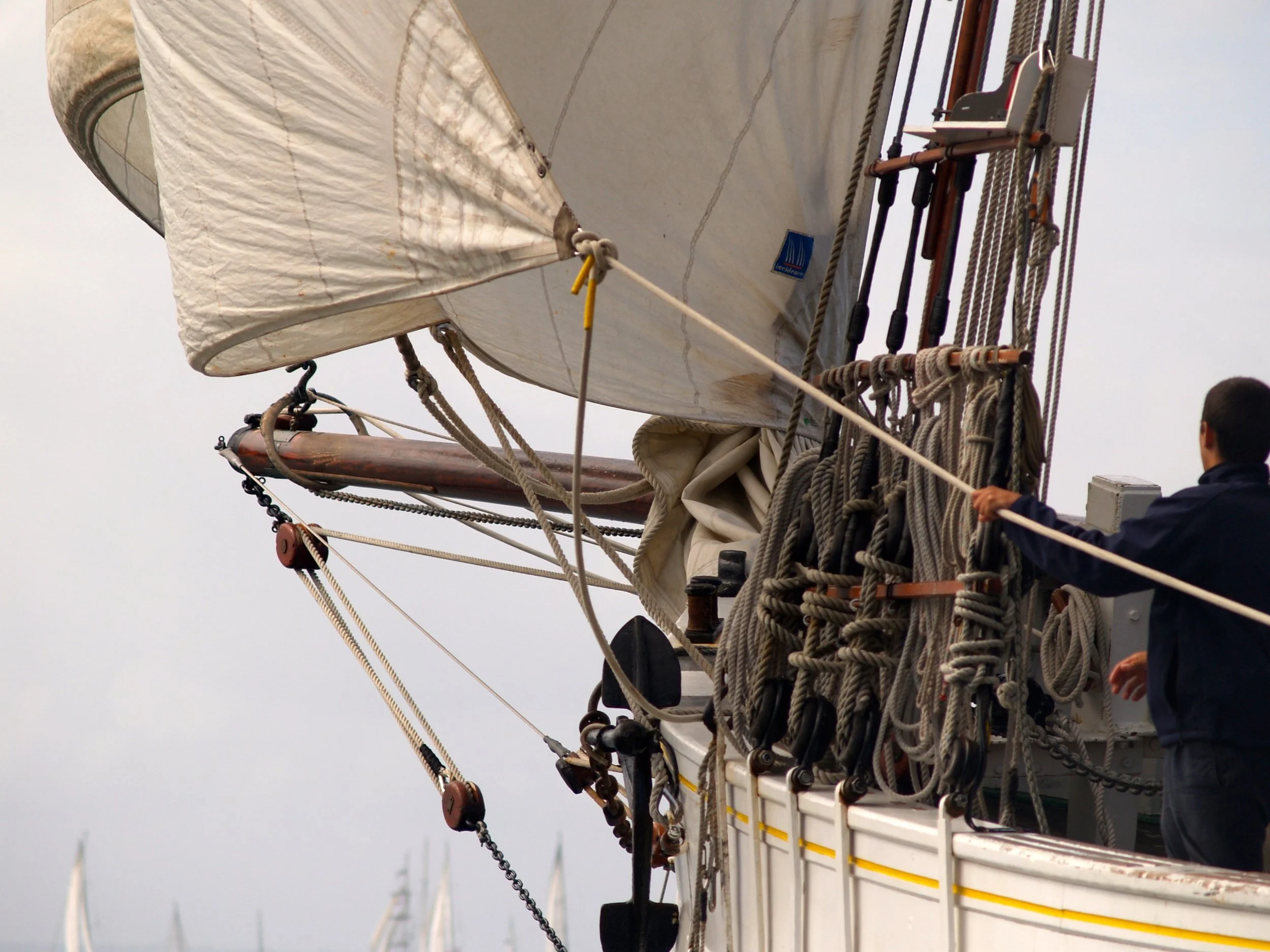 Le mât d'un voilier avec ses cordages, voiles enroulées, et une personne manipulant une ligne, vue à la proue d'un bateau en mer par temps nuageux.