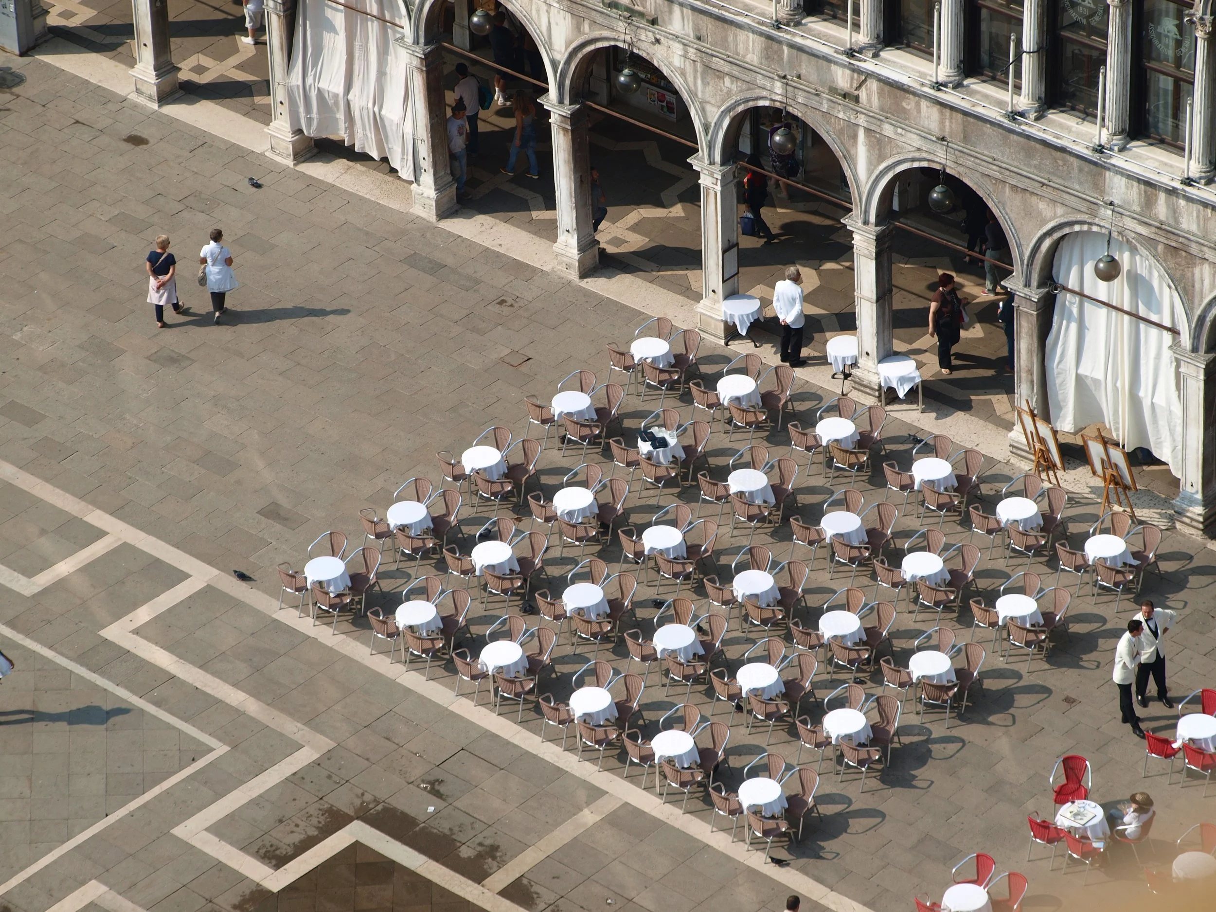 Vue aérienne d'une place pavée avec des tables à manger rondes avec des chaises en bois, une façade ancienne avec des arches et des lampes suspendues, des personnes marchant et se tenant dans la zone, certains en groupes et d'autres seuls.