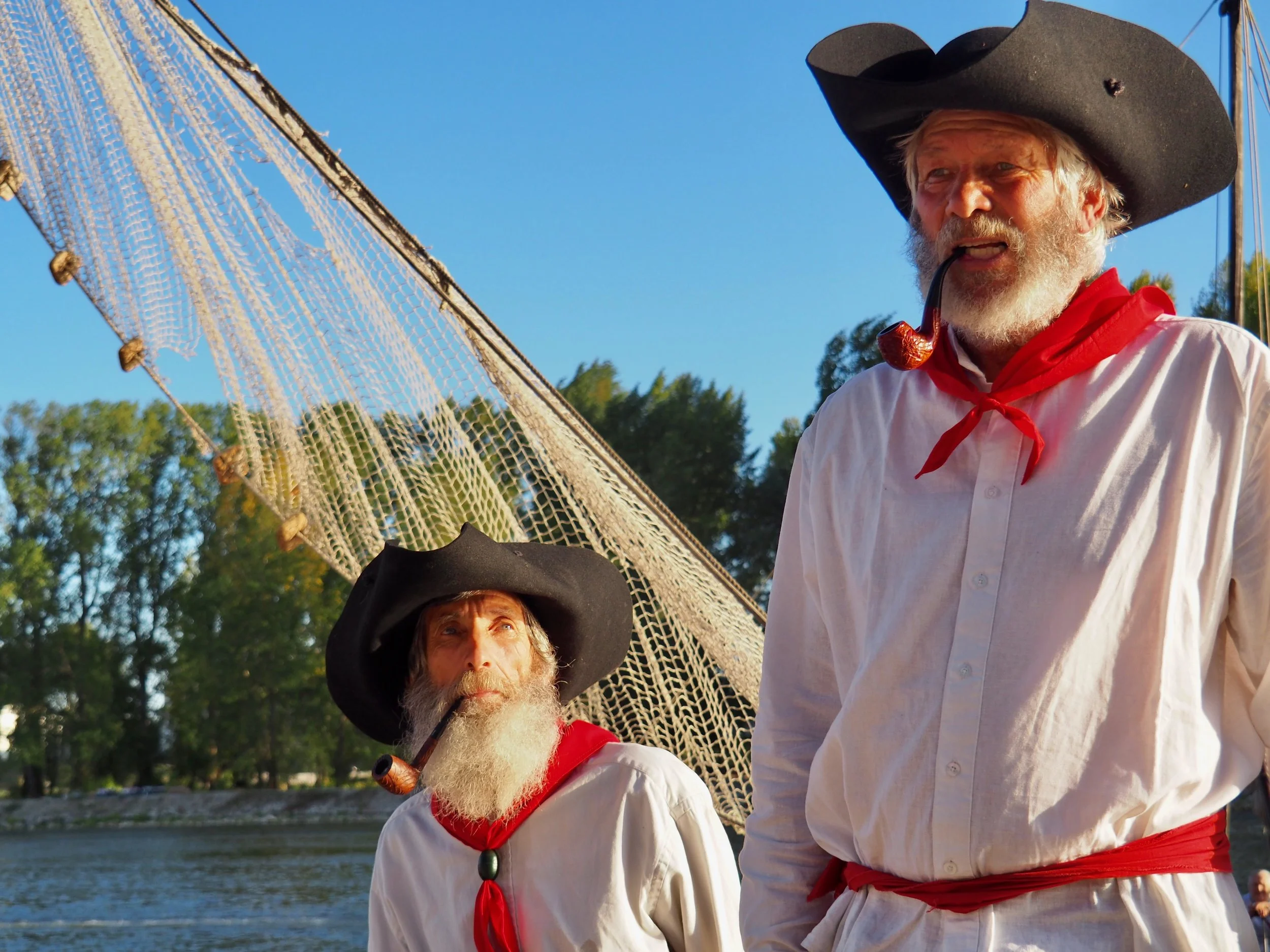 Deux hommes habillés en pirates avec des chapeaux noirs, des pipes, en costume blanc avec des foulards rouges, se tenant près d'une rivière sous un ciel bleu clair, un très grand navire en bois derrière eux.